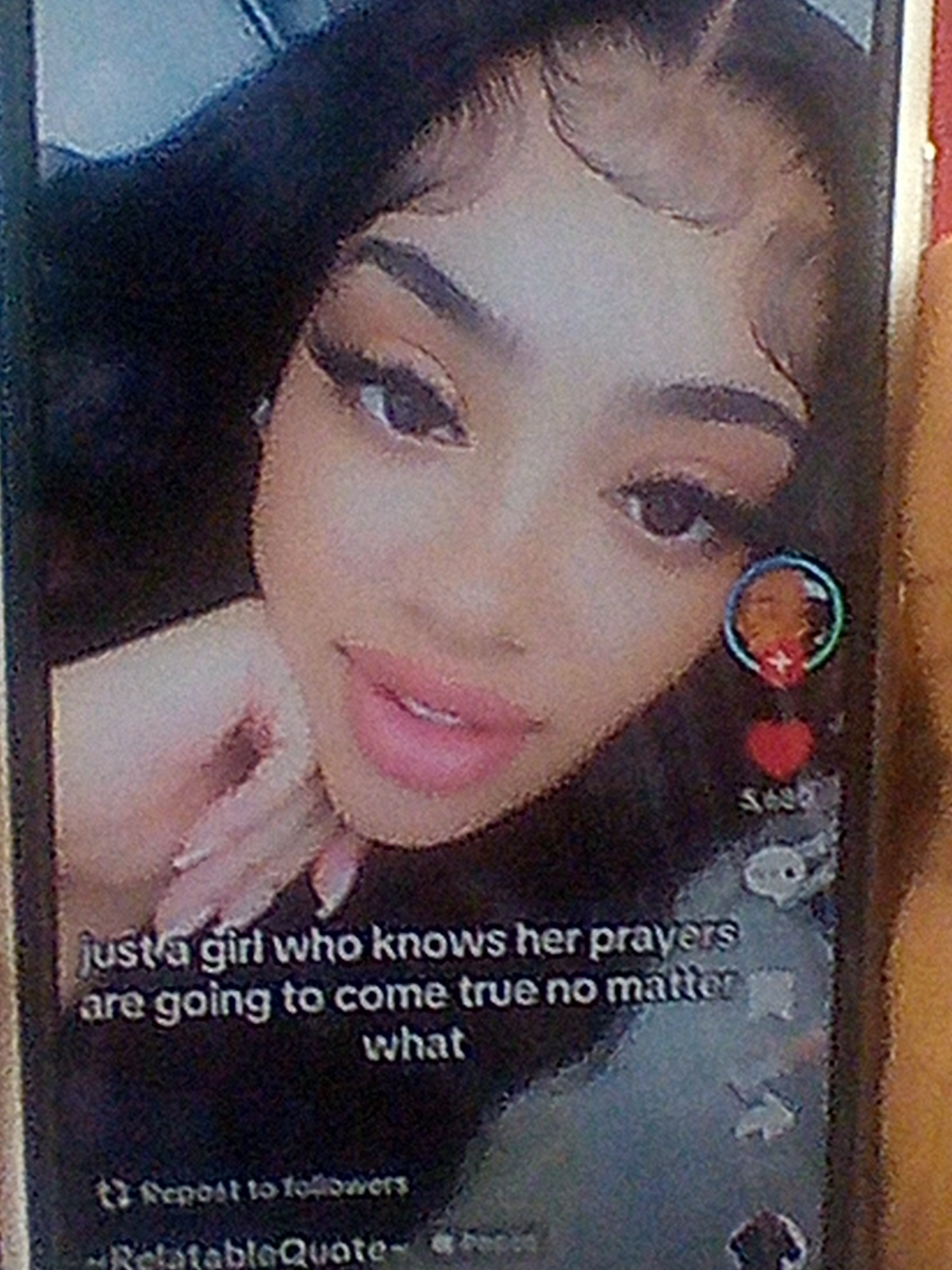 A close-up shot on a mobile device screen displays a portrait of a young woman with dark hair styled with baby hairs. She is looking towards the viewer with a soft expression, her lips slightly parted and painted with pink lipstick. Her eyes are emphasized with dark eyeliner and mascara, and her eyebrows are well-defined. A hand is visible near her face, with manicured fingernails.
Overlaying the image is text that reads: "just a girl who knows her prayers are going to come true no matter what". Below this, smaller text indicates "Repost to followers" and "-RelatableQuote-". On the right side of the screen, there are social media interaction icons, including a profile picture within a circle, a red heart icon with a number below it, and other smaller icons.
The image appears to be a screenshot or a photo of a screen, possibly from a social media platform like TikTok, given the interface elements. The background of the screen is dark, and the overall lighting suggests an indoor setting. There is no discernible information within the image that can be used to pinpoint its location in Bukuru, Nigeria.