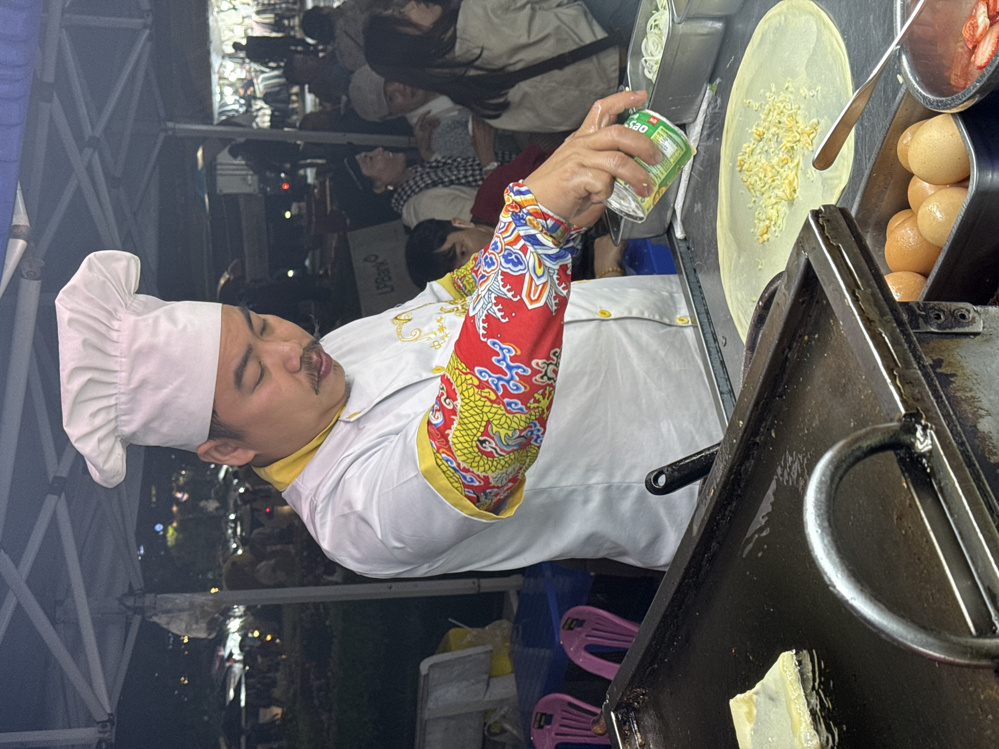 In Da Lat, Vietnam, a street food vendor wearing a white chef's hat and a white uniform with red and yellow dragon-embroidered sleeves prepares food at a food stall. He holds a can of ingredients over a thin crepe on a flat cooking surface, sprinkling the contents onto it. Next to the crepe are containers holding ingredients like eggs and strawberries. The food stall is set up outdoors, with a crowd of people in the background and chairs nearby. The scene is lit by overhead lights.
