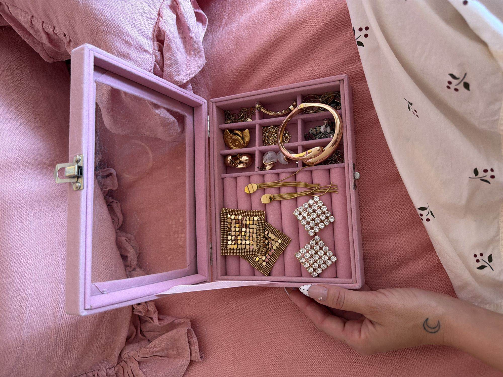 A pink jewelry box filled with various pieces of jewelry sits on a pink bedspread. The box is open, revealing earrings, necklaces, and bracelets. A person's hand with a crescent moon tattoo on the wrist is visible near the bottom right of the frame. The scene is located in Mengwi, Indonesia.