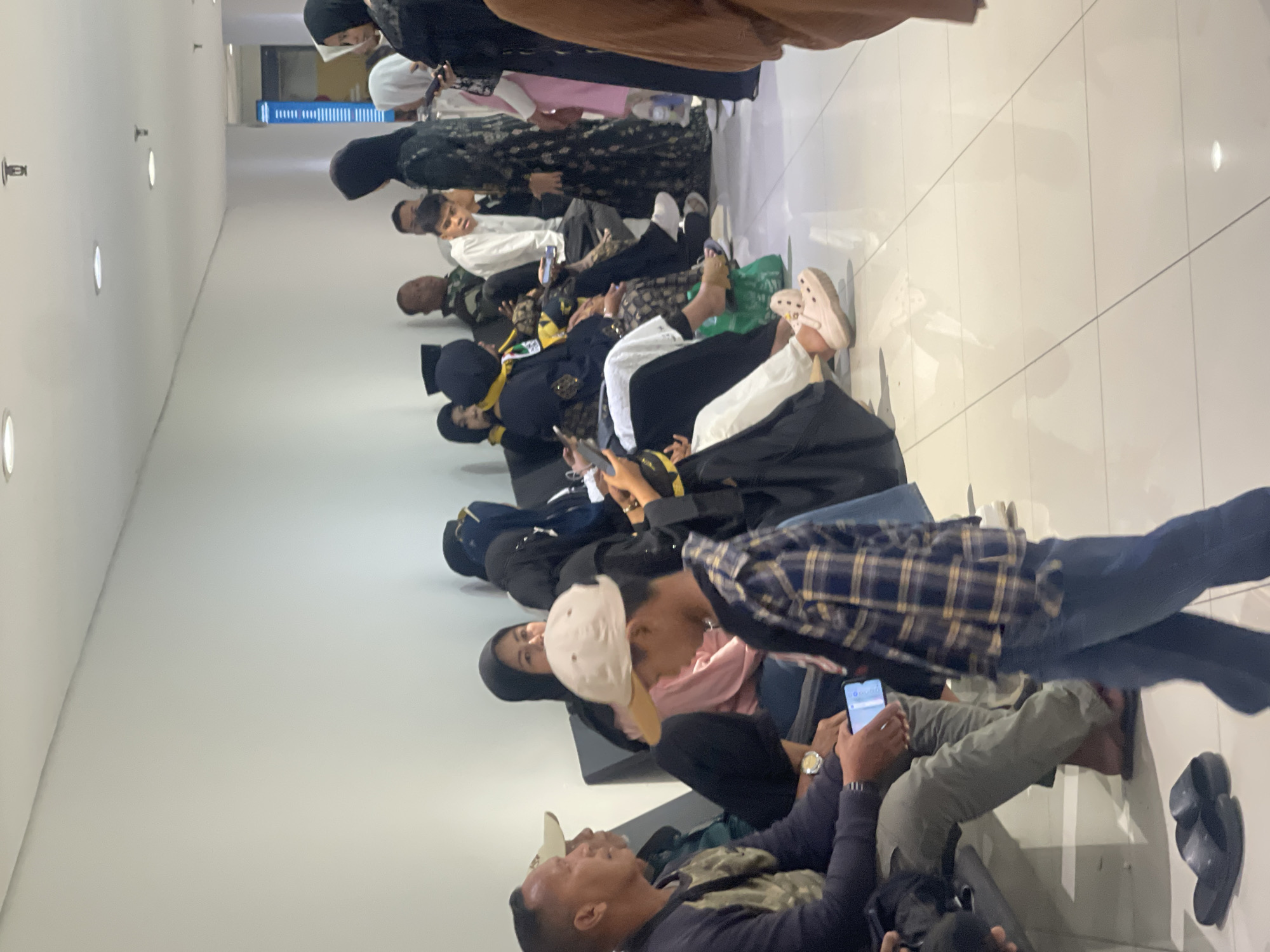The image depicts a waiting area with multiple individuals seated on a long bench and standing. The environment appears to be indoors with white walls and a tiled floor, suggesting a public space such as an airport, train station, or clinic. Several people are wearing traditional Islamic attire, including hijabs and long dresses. One person in the foreground is a young boy wearing a plaid shirt and a light-colored baseball cap, looking down at a mobile phone. An adult male to his left is also looking at a phone. Another individual, a woman in a black hijab, is visible in the background, as are other individuals in the process of waiting. The scene conveys a sense of waiting or transit. The provided hint about the location being in Tangerang, Indonesia, suggests this might be a transit hub or public facility within that city.