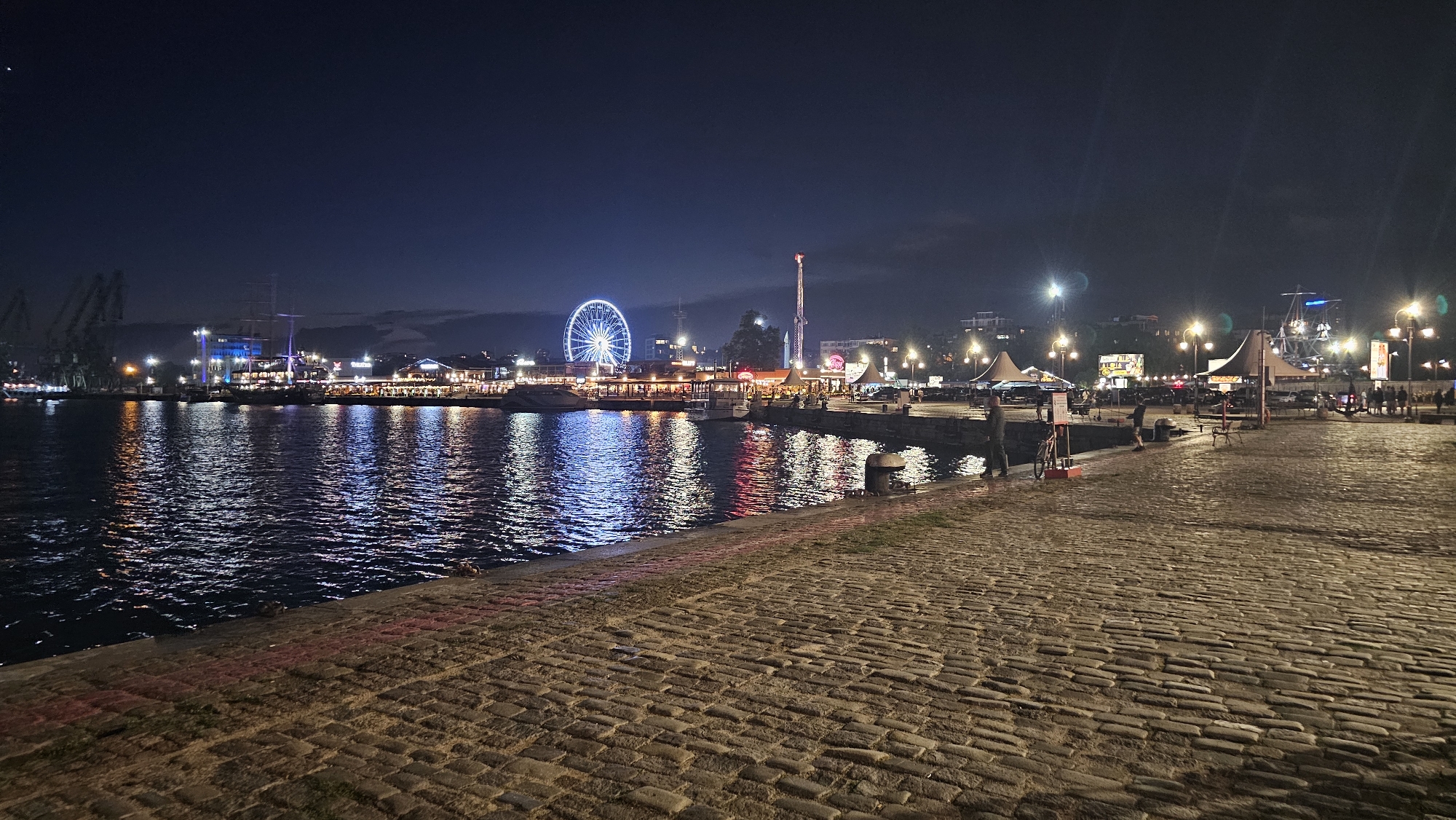 The image captures a nighttime view of the Varna Sea Garden, a waterfront promenade in Varna, Bulgaria. In the foreground, a cobblestone walkway stretches along the edge of the water. The Black Sea reflects the vibrant lights from the opposite shore, which is lined with buildings, restaurants, and amusement park attractions.
Prominently featured is a large Ferris wheel, illuminated with bright white lights. To its right, a tall tower, possibly a ride or a landmark, is also lit up. Several tents and smaller structures are visible along the waterfront, suggesting market stalls or dining areas. Various boats are moored in the harbor, and a large sailing ship is docked further down the quay. Cranes are visible in the distance, indicating a port area.
Several individuals are scattered throughout the scene, some walking along the promenade, others gathered near the waterfront establishments. One person is seen with a bicycle. The overall atmosphere is lively and illuminated, typical of a popular evening destination. The sky is dark, with some faint clouds visible.