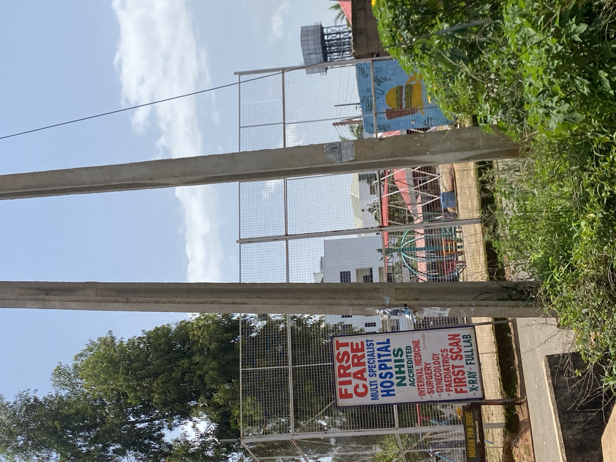 A vertical outdoor shot, taken from a slightly low angle, shows a "FIRST CARE MULTI SPECIALIST HOSPITAL NHIS ACCREDITED" sign. The sign is white with red, blue, and black text, and it lists services such as "INTERNAL MEDICINE", "SURGERY", "GYNECOLOGY", "PAEDIATRICS", "FIRST SCAN", "X-RAY", and "FULL LAB". The sign is attached to a chain-link fence, behind which is a playground with blue and red structures. In the background, a multi-story white building is visible, along with a water tower and a building with a mural of a hamburger. Two large concrete poles, one in the foreground and one slightly behind it, partially obscure the view of the background. Trees and green foliage are also present in the image. The sky is blue with scattered white clouds. A street sign reads "MARIAN PAM DRIVE". The location is the city of Bar Jirgi Summa, Nigeria.