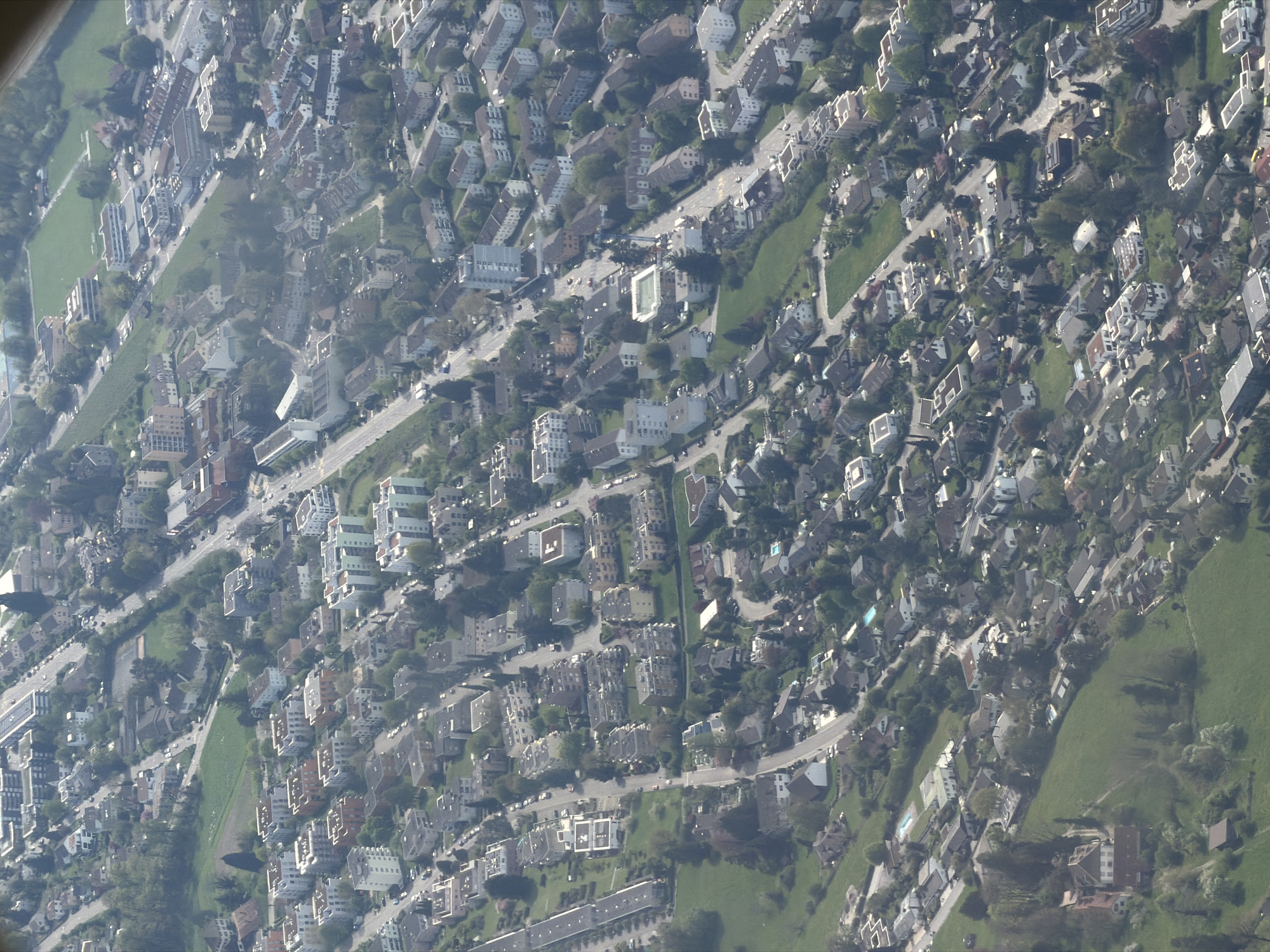This aerial image captures a sprawling urban landscape, likely a residential area, viewed from a high vantage point, possibly an aircraft or drone. The scene is dominated by a dense network of buildings, roads, and interspersed green spaces.
The subjects primarily consist of numerous multi-story buildings and individual houses, varying in architectural style but generally featuring light-colored facades and a mix of flat or sloped roofs. Several buildings appear to be modern apartment complexes, some with visible swimming pools in courtyards or on rooftops. A comprehensive system of grey asphalt roads winds through the area, with white road markings visible on some thoroughfares. Numerous small, dark-colored cars are seen parked along streets and in parking areas, indicating active use of the infrastructure.
Significant green areas are present, ranging from well-maintained lawns and gardens surrounding buildings to larger, undeveloped grassy fields and clusters of trees. These green spaces break up the urban density.
No specific activity or event is clearly discernible due to the high altitude, other than the general flow of traffic suggested by the presence of vehicles.
Notable details include the bright daylight, with shadows cast by buildings and trees suggesting either a morning or late afternoon sun angle. The overall visibility is good, implying clear weather conditions. There is no visible text in the image. While the architecture and urban layout are consistent with a European city, there are no specific landmarks within the image to definitively confirm the provided context of Sofia, Bulgaria.