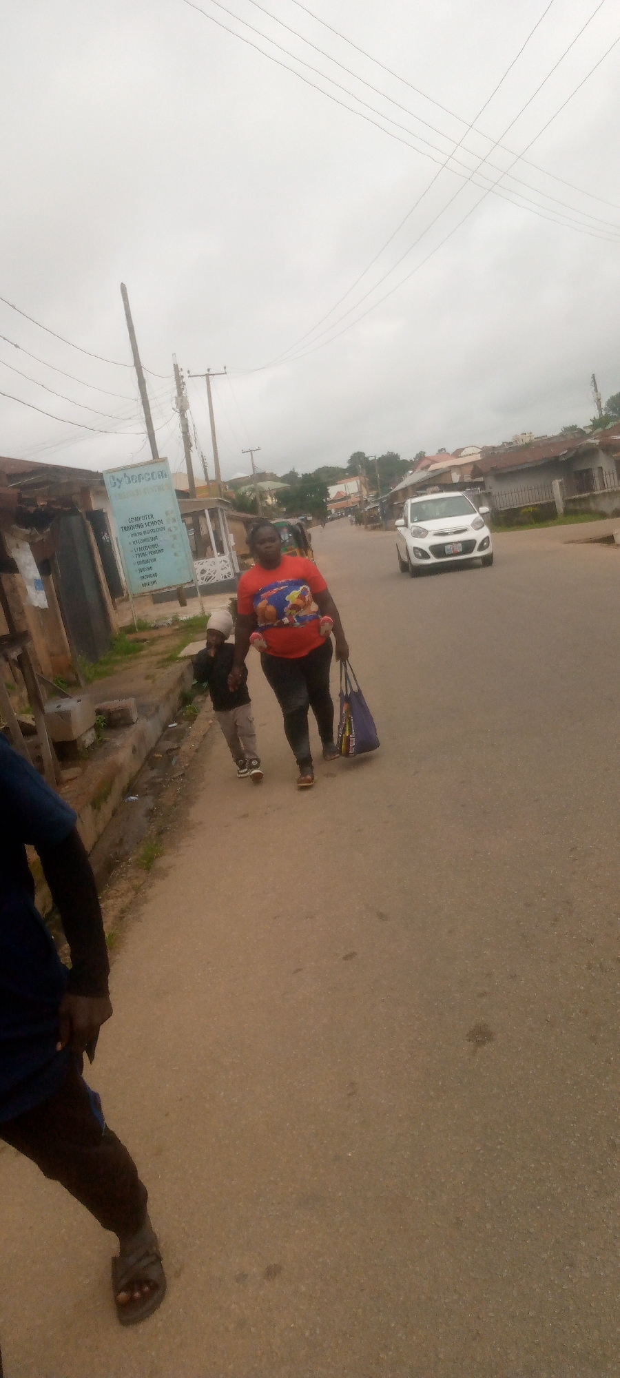 A woman and a child are walking down a street in an urban area. The woman is wearing a red t-shirt with a graphic on it and dark leggings. She is carrying a blue and purple tote bag. The child is wearing a light-colored beanie, a dark jacket, and light-colored pants. They are walking on a paved road with buildings and utility poles on either side. The sky is overcast. A white car is visible driving in the distance. Another person is partially visible in the foreground on the left side of the image.