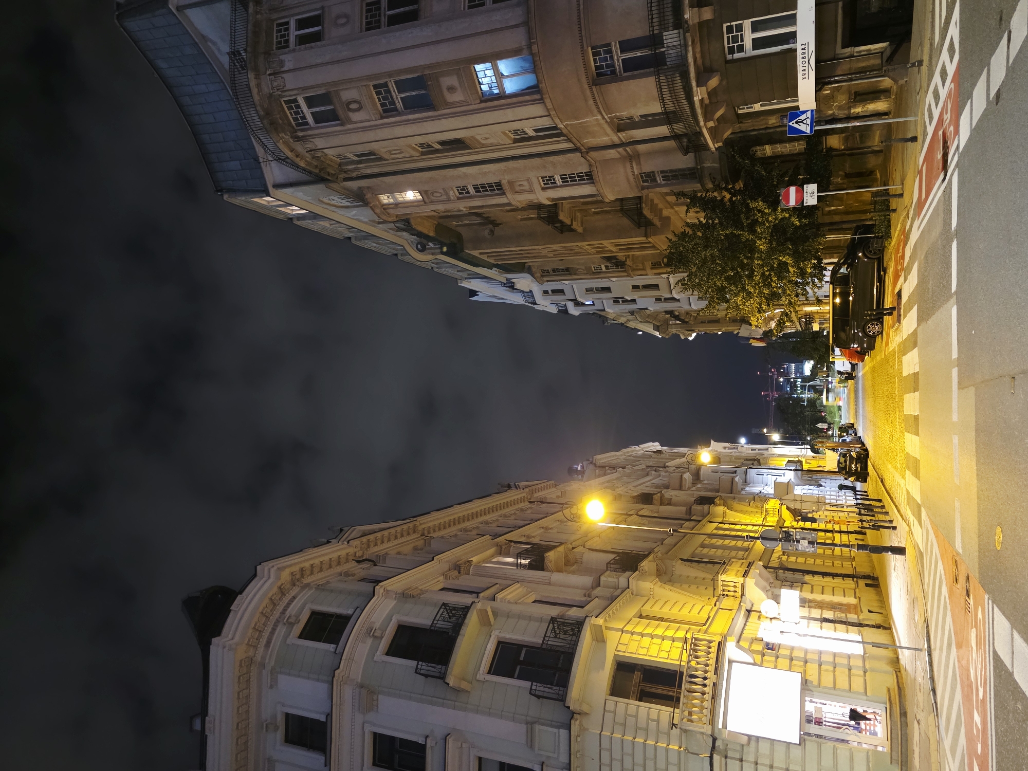 The image captures a street scene at night in Warsaw, Poland. On the left, a pale yellow, ornate building with multiple stories and arched windows is illuminated by streetlights. The building features decorative balconies with intricate metal railings and detailed architectural elements. To the right, a similar-style building, rendered in shades of beige and gray, stands tall. This building also displays balconies and a curved facade, with lights visible in some of its windows.
The street itself is paved with cobblestones, and prominent crosswalks with white and red markings are visible. Several cars are parked along the right side of the street, and a street lamp with a bright yellow bulb casts a strong glow on the pavement. In the background, the night sky is filled with dark, cloudy formations. Further down the street, hints of other buildings and some construction cranes are discernible, suggesting an urban environment. The overall impression is of a well-lit, historic city street at night.