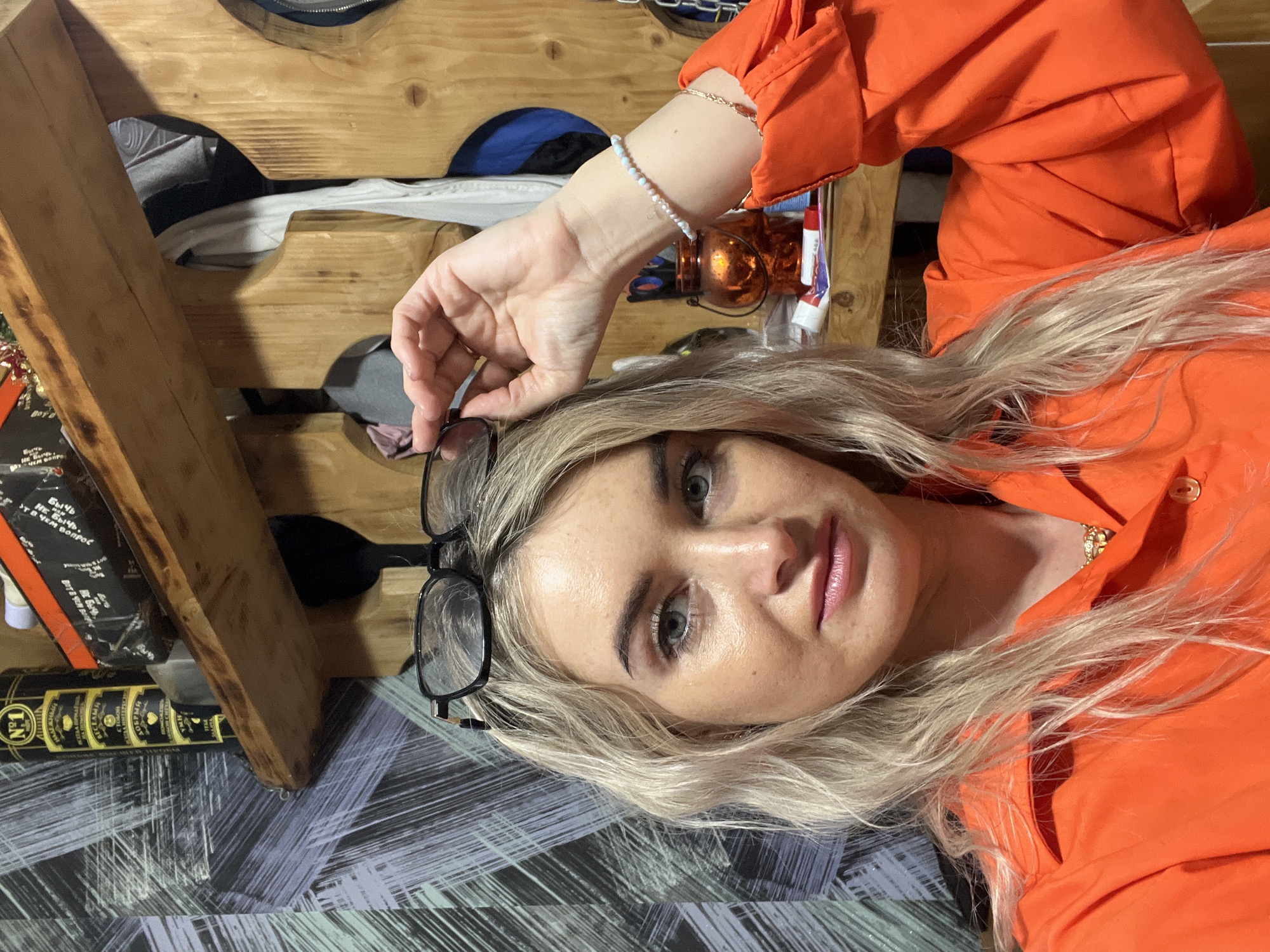 The image shows a fair-skinned woman with blonde hair, wearing an orange shirt, lying down. She has glasses in her hand. A wooden structure is partially visible to the left, with items stored in it. There is a bottle with gold lettering also on the left. The woman wears a delicate beaded bracelet on her left wrist. The surface she lies on has a patterned design in shades of grey and blue. There are no indicators in the image identifying a specific location, or the city of Chaikovsky.