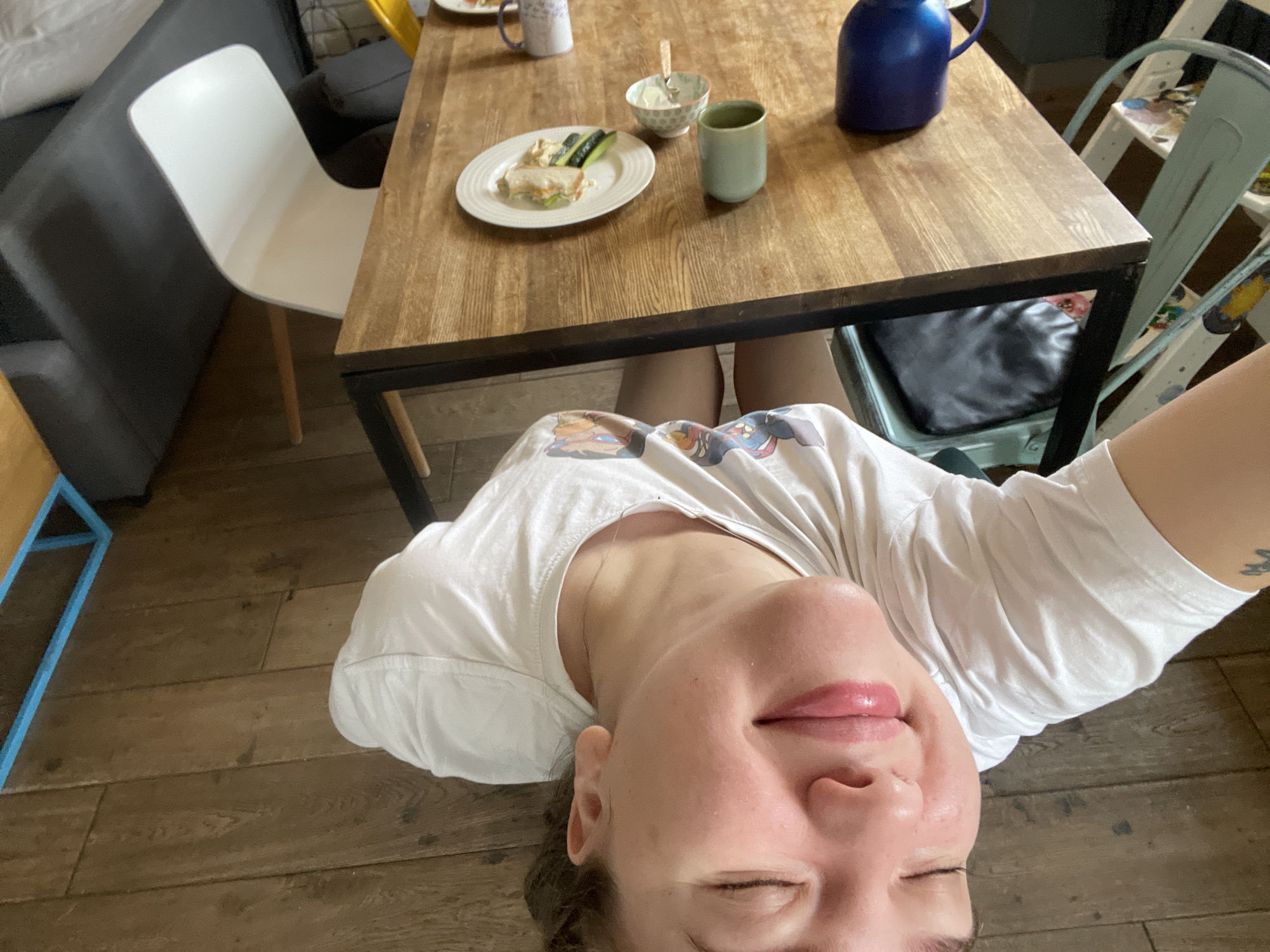 The image shows a person in a white t-shirt at a table with food and beverages. A plate contains a sandwich and cucumbers. Other items on the table include a blue pitcher, a cup, and a small bowl. There are chairs around the table, and the floor is made of wood. The location cannot be determined.