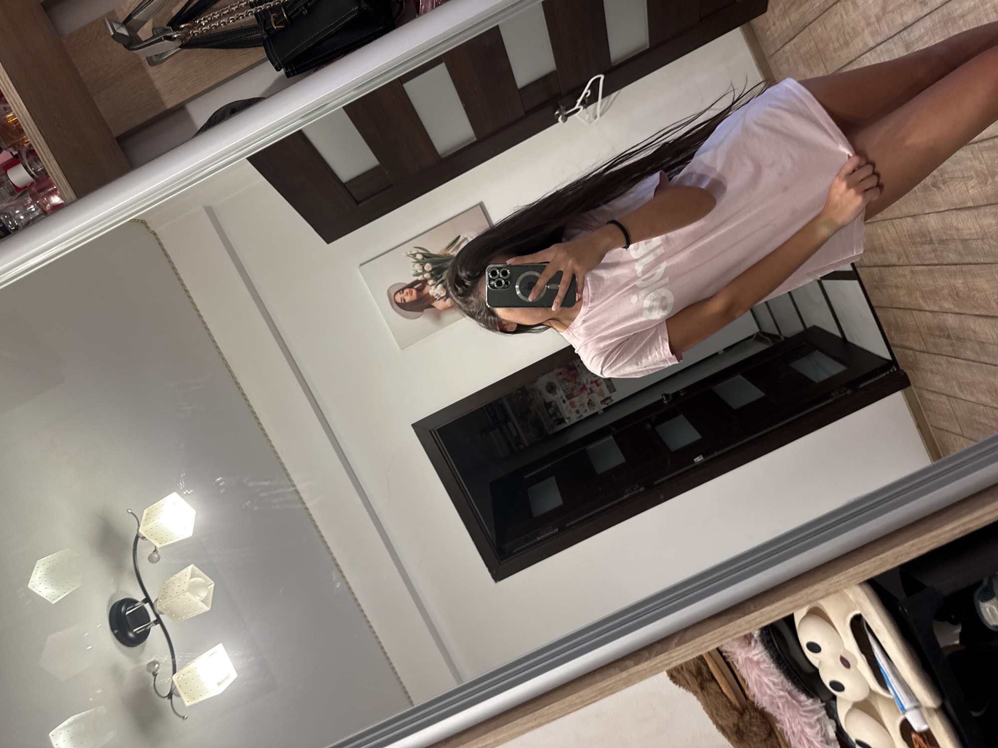 A young woman with long, dark hair is taking a mirror selfie in a room. She is wearing a pink t-shirt. The room has a wooden floor, a dark wooden door, and a painting on the wall of a woman wearing a hat and holding flowers. In the foreground, part of a closet or shelving unit is visible, with a plush toy or slippers on display. A ceiling light fixture with three lamps is reflected in the mirror. The setting appears to be residential.