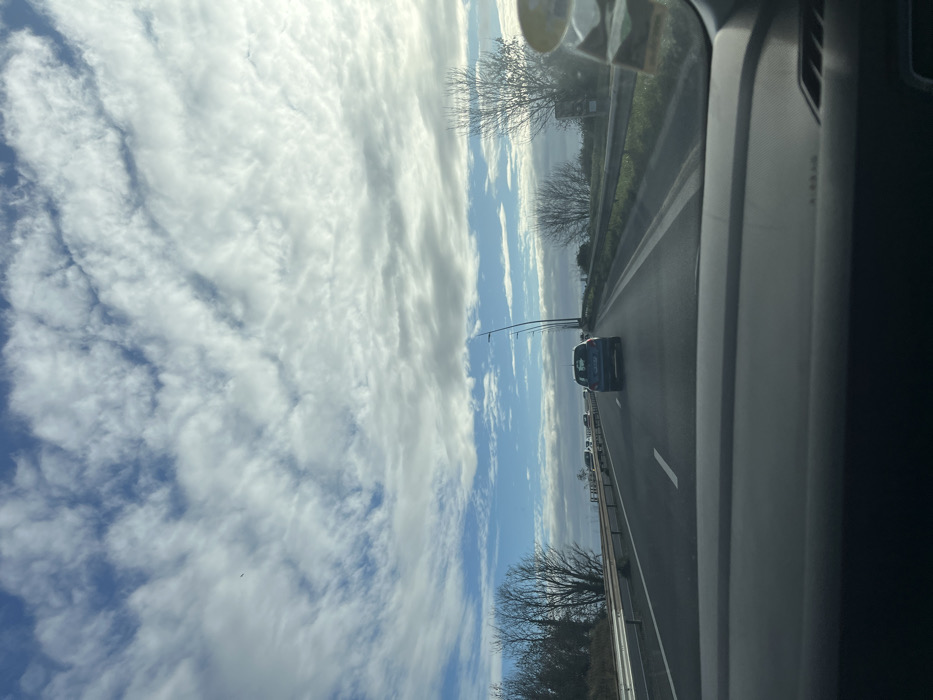 The image depicts a daytime view from the interior of a vehicle on a multi-lane highway in Hérouville-Saint-Clair, France. The foreground shows the top portion of a grey vehicle dashboard. The asphalt roadway, marked with white solid and dashed lines, extends into the distance. A dark-colored hatchback, possibly a Renault Clio, is positioned directly ahead in the rightmost lane, with additional vehicles visible further down the road. Metal guardrails line both sides of the highway. The left roadside features a sloped embankment with bare deciduous trees and some evergreen vegetation. The right roadside exhibits bare deciduous trees, a series of streetlights with curved black poles, and a partially visible roadside sign. The sky above is partly cloudy, displaying patches of blue sky interspersed with widespread white and grey cloud formations. No individuals are visible in the scene.