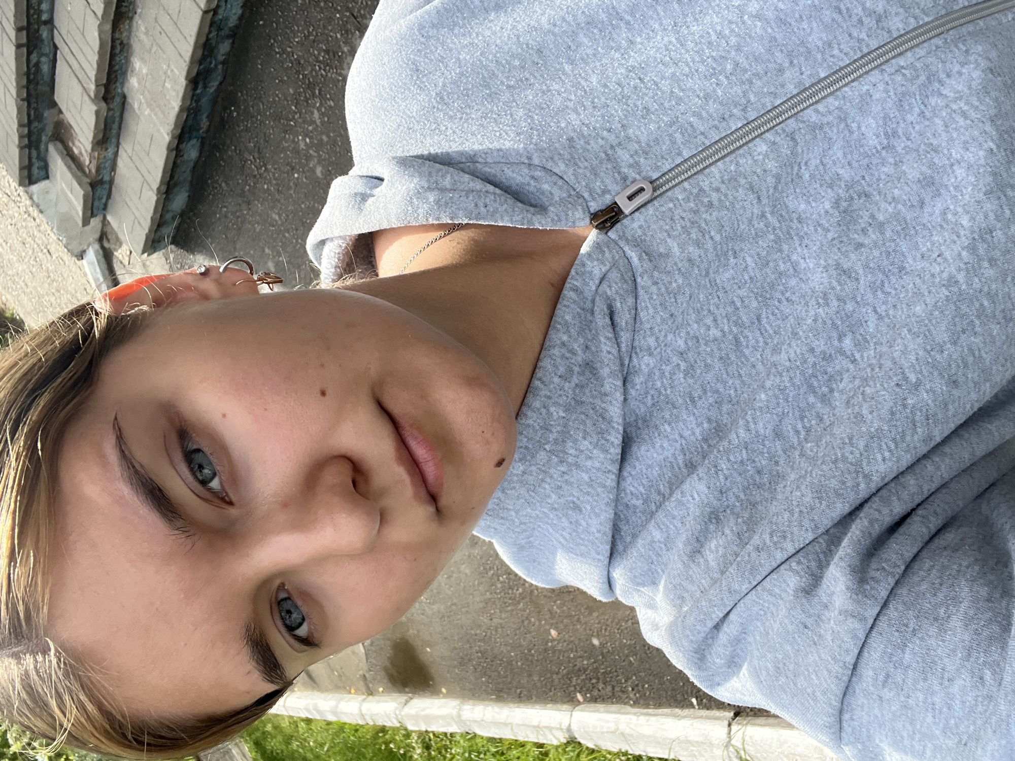 A close-up selfie shows a young woman with light brown hair and blue eyes, wearing a light gray hooded jacket. She has several piercings in her left ear. Behind her, there are outdoor stairs and a paved area. The lighting suggests it is daytime. The image was taken on a paved area next to a grassy strip, with concrete stairs in the background. The scene appears to be outdoors in a residential or public area. The exact location in Chaikovsky, Russia, cannot be determined from the image alone.