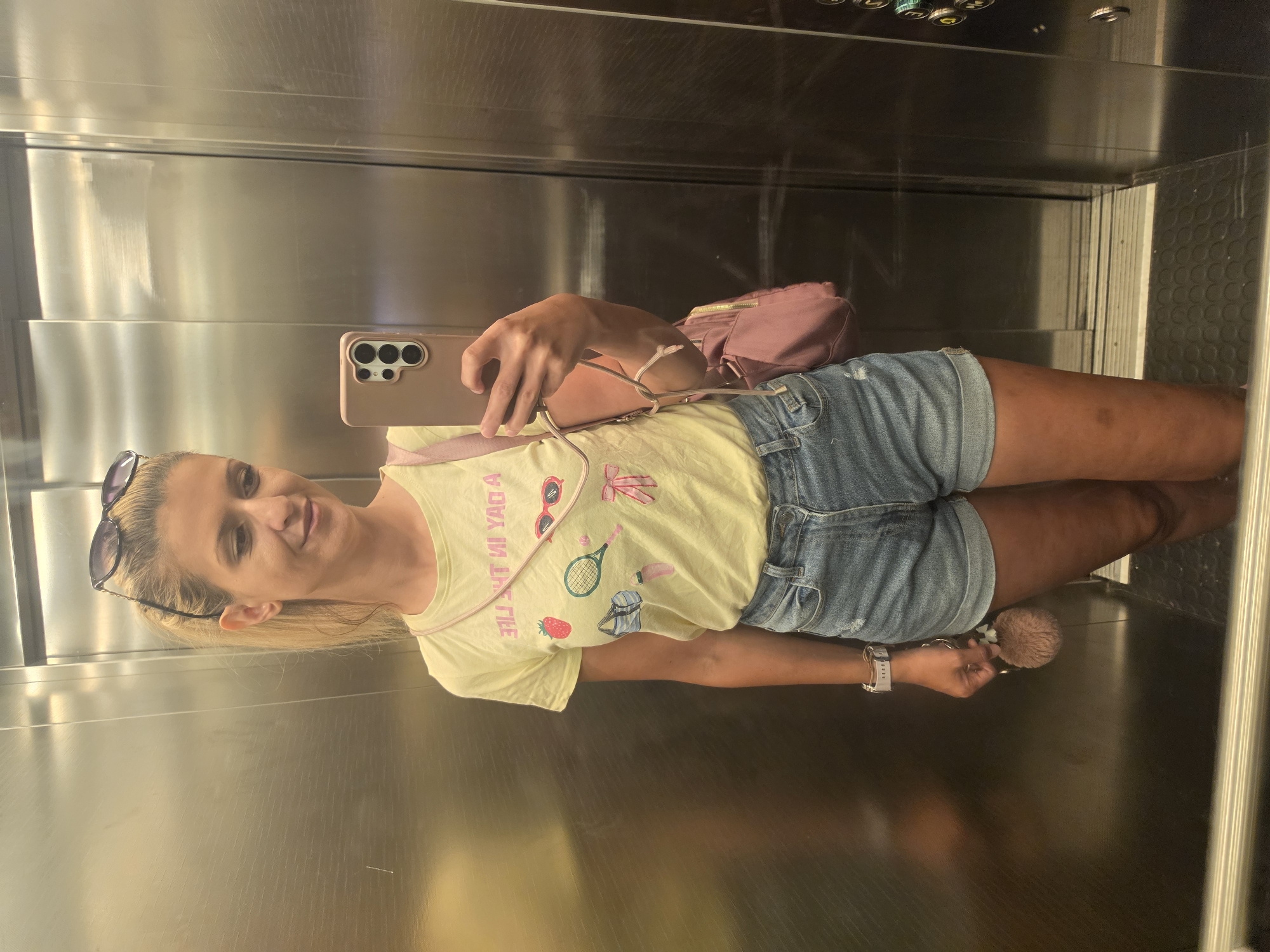 A young Caucasian woman with blonde hair pulled into a ponytail is standing in an elevator. She is wearing a yellow t-shirt with graphics, denim shorts, and a pink crossbody bag. She is holding a mobile phone in her right hand, taking a selfie. Sunglasses are resting on top of her head. A fluffy keychain dangles from her left hand. The elevator walls are made of brushed metal.