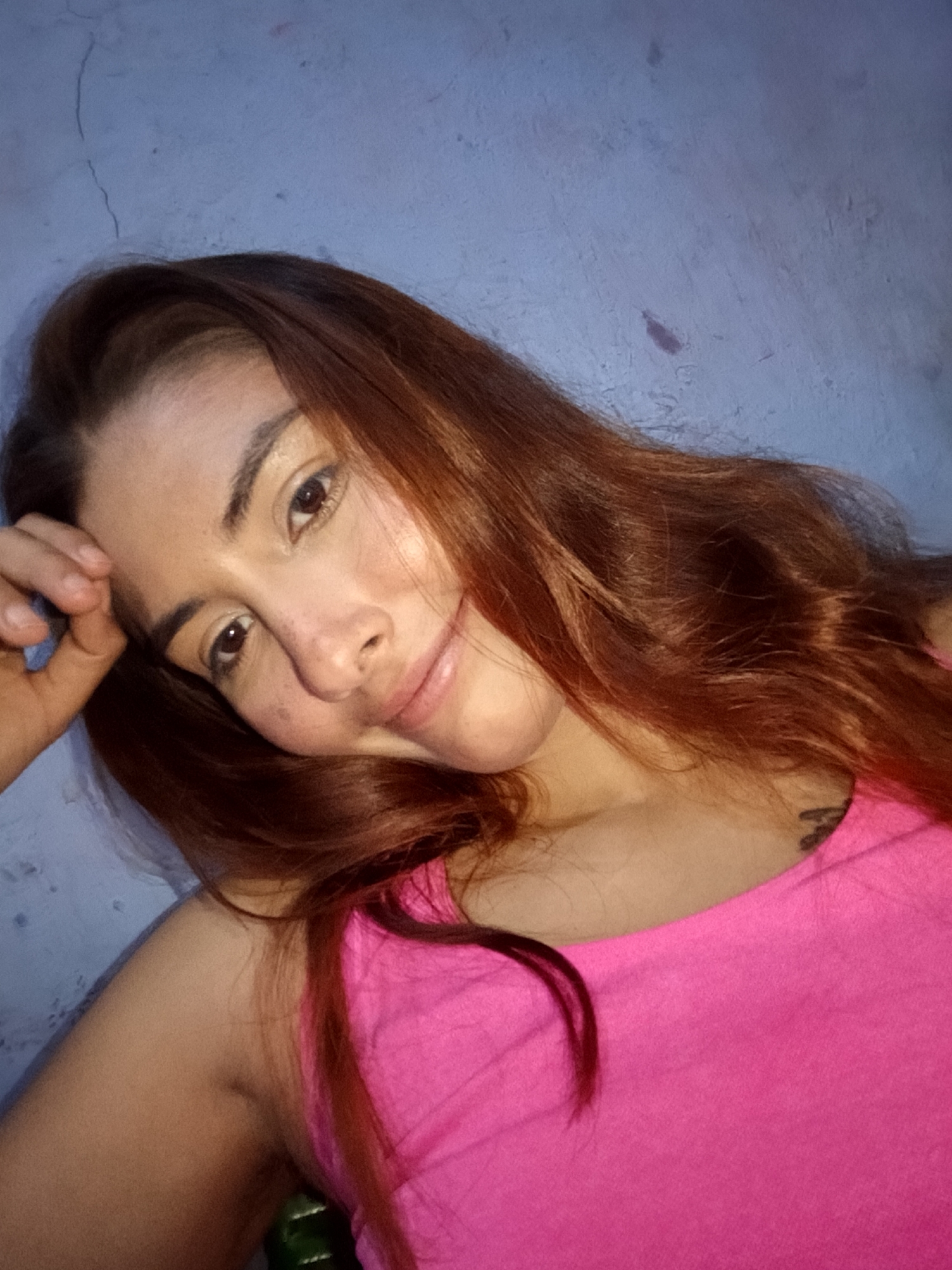A young woman with auburn hair and light skin is shown in a selfie taken indoors against a pale blue, textured wall. She is wearing a bright pink tank top. Her left hand is resting on her forehead, and her head is tilted. She has dark eyes and a slight smile. There appears to be a tattoo on her upper left shoulder. The lighting suggests it may be evening or indoors with artificial light. The location is identified as Juan Griego, Venezuela, although no specific landmarks or contextual clues in the image confirm this.