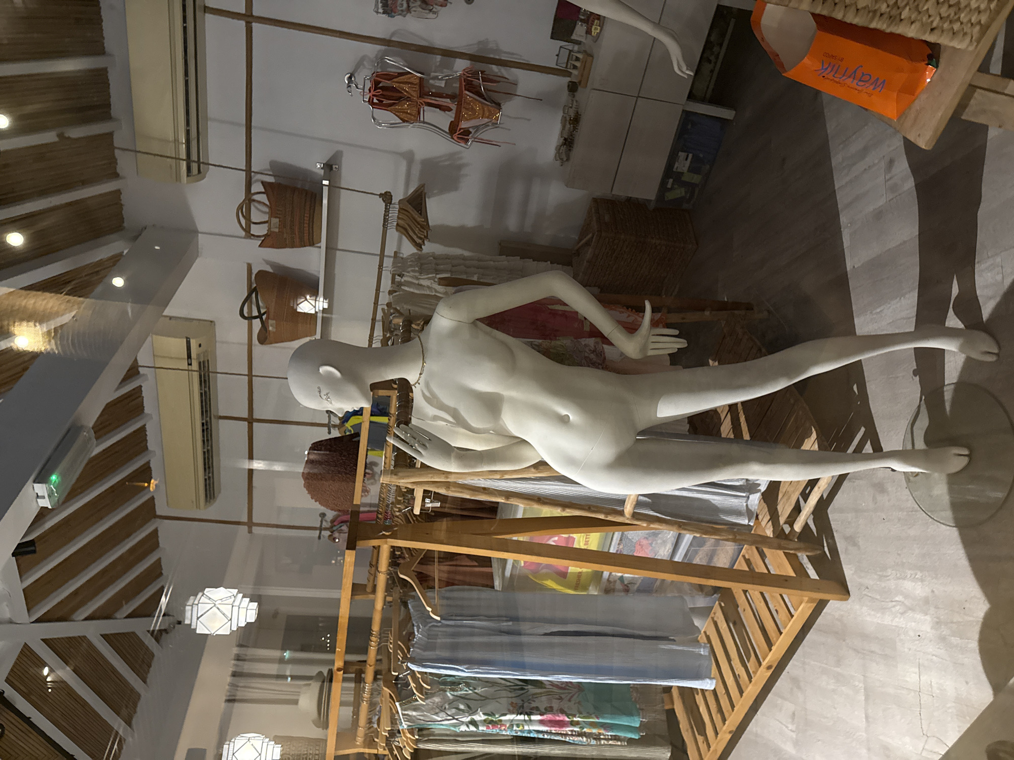 A retail store interior is depicted, likely a boutique specializing in women's apparel. The setting appears to be in Dubai, United Arab Emirates, given the presence of a prominent orange shopping bag with Arabic text and branding. The store features a rustic-chic aesthetic with exposed wooden beams in the ceiling, bamboo-like clothing racks, and woven storage baskets.
Dominating the foreground is a full-body female mannequin, posed in a three-quarter view, facing towards the left of the frame. The mannequin is a pale, matte white and is dressed in a minimal, light-colored garment that accentuates its form. It has a delicate necklace. Its left hand is raised, with the index and middle fingers extended, as if gesturing or holding an invisible object, while its right hand rests on its hip.
Behind the mannequin, a wooden clothing rack displays various garments, including dresses with floral patterns and solid-colored trousers. A large, clear plastic bin with some yellow packaging is visible beneath the clothing rack. Further in the background, more clothing racks are present, along with woven baskets on shelves and decorative lighting fixtures. A separate display to the right features a mannequin form showcasing a shimmering orange bikini.
The store's environment is lit by a combination of overhead lighting, including modern geometric pendant lights and recessed spotlights, and natural light, possibly from a storefront window. The floor is a light-colored, textured material, possibly concrete or polished stone. The overall atmosphere suggests a relaxed, possibly beach-themed or resort wear boutique.