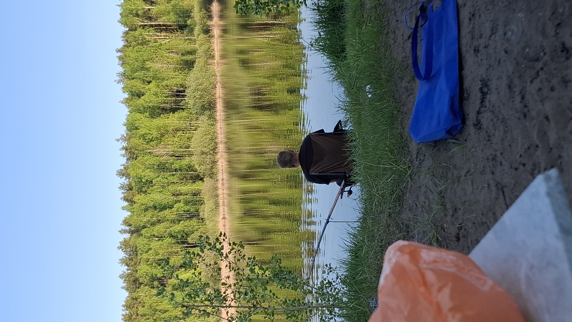 A man is fishing on a body of water, likely a lake or river, in Chaikovsky, Russia. The shoreline is grassy with some muddy patches. A blue tote bag sits on the ground near the man. The lake reflects the treeline on the opposite shore. A fishing rod extends into the water from the man's location. Orange packaging is visible on a surface near the bottom of the frame. The sky is visible and clear. The photo is taken outdoors during daylight.