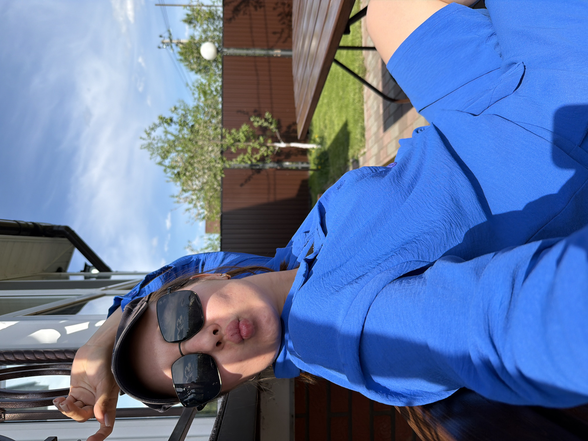 A person is photographed in an outdoor setting. The person is wearing a blue shirt and sunglasses, with one hand resting on a dark surface. The person is looking at the camera. In the background, there is a wooden table, a green lawn, a fence, and a tree under a bright, partly cloudy sky. The location is Chaikovsky, Russia.