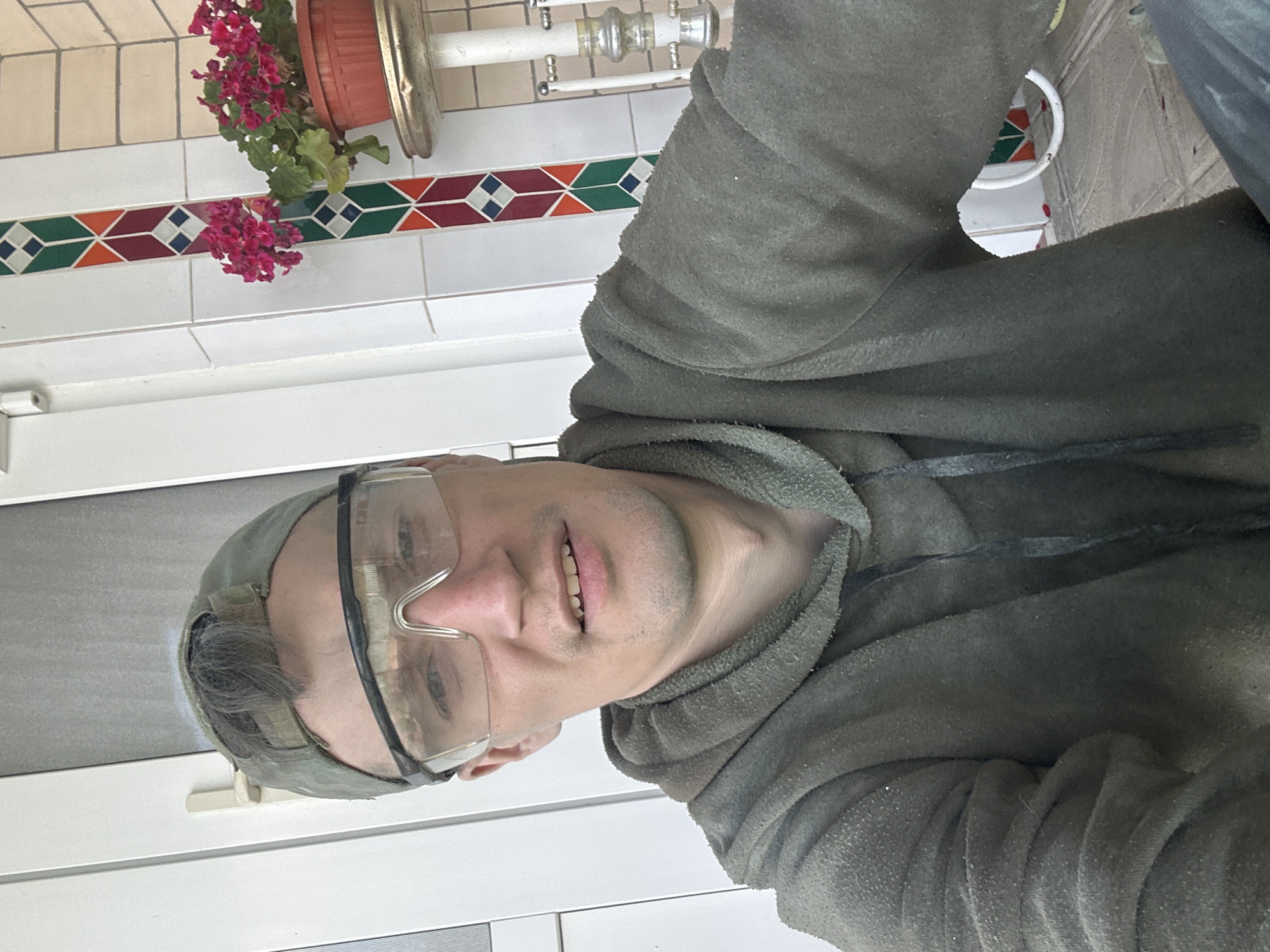 The image shows a close-up, selfie-style shot of a person wearing safety glasses and a green hooded jacket. The person has short, dark hair visible under a green baseball cap and appears to be smiling. The background includes a tiled wall with a decorative border featuring geometric patterns in red, green, and blue. A plant with pink flowers in a terracotta pot is also visible, hanging slightly to the left. In the upper right corner of the image, parts of some metal fixtures, possibly plumbing or light fixtures, are visible. The overall impression is that the person is in an indoor or outdoor area with tiling and some decorative elements. The image appears to be taken during the daytime, and the location context provided indicates Makiv, Ukraine.