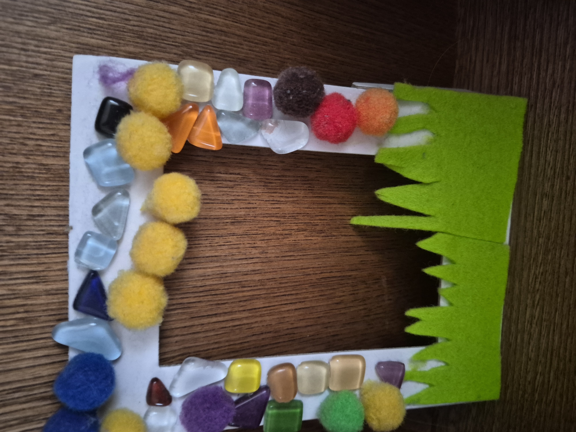 A handmade picture frame decorated with colorful pom-poms and polished stones is displayed on a wooden surface. The frame itself is made of white cardboard, with a rectangular opening in the center. The border of the frame is adorned with an assortment of small, round, fuzzy pom-poms in yellow, blue, purple, green, red, and brown. Interspersed among the pom-poms are various shapes and colors of polished glass or stone pieces, including clear, light blue, dark blue, purple, orange, and amber. The bottom of the frame features a green felt cutout resembling grass with pointed tips. The background appears to be a wooden cabinet or wall with vertical grain. The lighting is soft, creating some shadows. The image is taken from a slightly elevated angle.