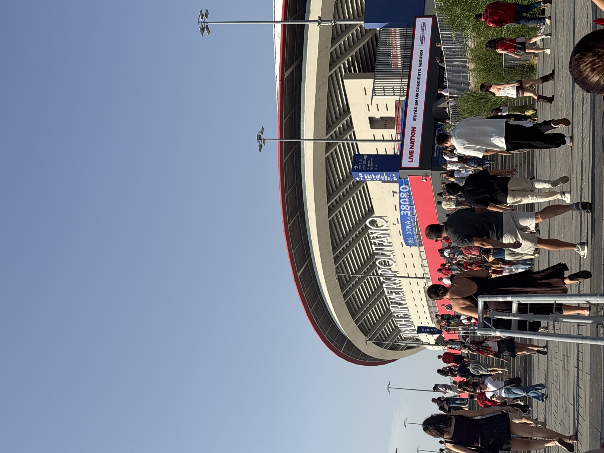 The image depicts the exterior of the Cívitas Metropolitano stadium in Madrid, Spain, on a clear, sunny day. A crowd of people are walking up a wide, paved staircase towards the stadium entrance. The stadium features a distinctive red roof and white architectural elements with large text reading "RYADH AIR METROPOLITANO". A large digital billboard displays "LIVE NATION ¡ESTÁS EN UN CONCIERTO SEGURO! RESPECT NATION". People are dressed in casual summer clothing, and many appear to be attending an event. The overall atmosphere suggests a gathering for a concert or sporting event.