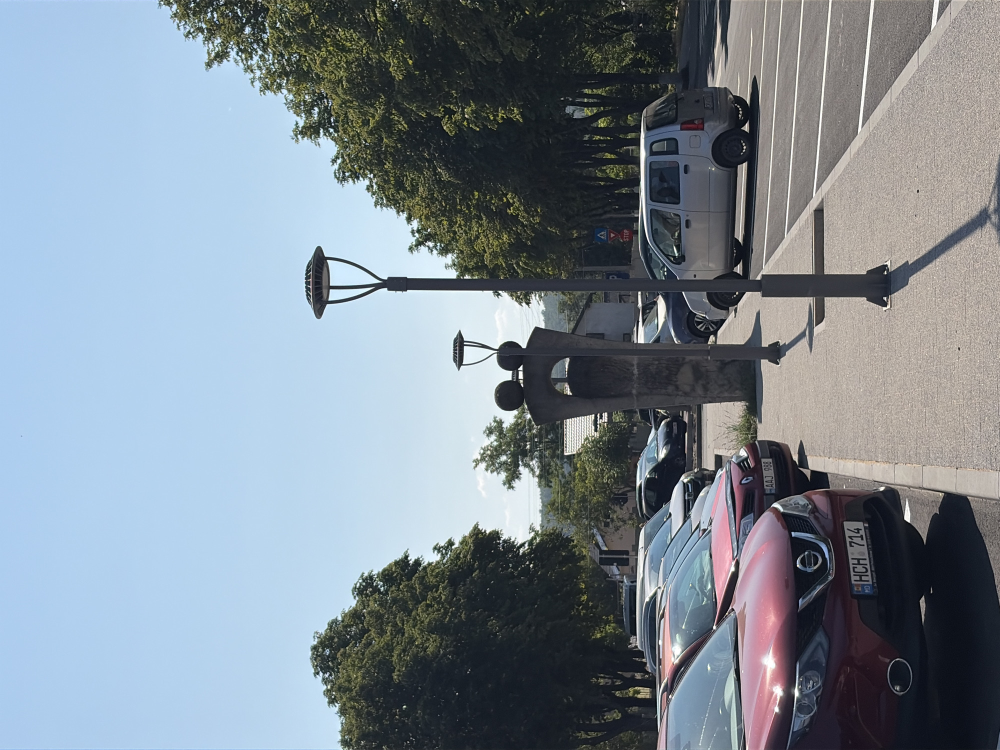 A clear, bright sky dominates the top half of the image. The lower half depicts a row of parked cars on a sunny day in what appears to be a public parking lot in Criuleni, Moldova. In the foreground, a metallic red Nissan Qashqai is parked facing left, with its license plate clearly visible: "HCH 714". Behind it, a red Renault Megane is parked, its license plate reading "AAJ 988". Further back, several other cars are parked in a line, including a silver Suzuki Wagon R. A tall, modern street lamp with a distinctive umbrella-shaped top casts a long shadow across the paved ground. To the left of the street lamp, a concrete sculpture or monument stands, with two spherical elements on top and a rough-hewn base. Trees with dense green foliage line the background, some reaching towards the sky. In the distance, buildings with tiled roofs are visible. A blue and white pedestrian crossing sign is also present, accompanied by a red stop sign. The scene is orderly, suggesting a typical urban environment during daylight hours.