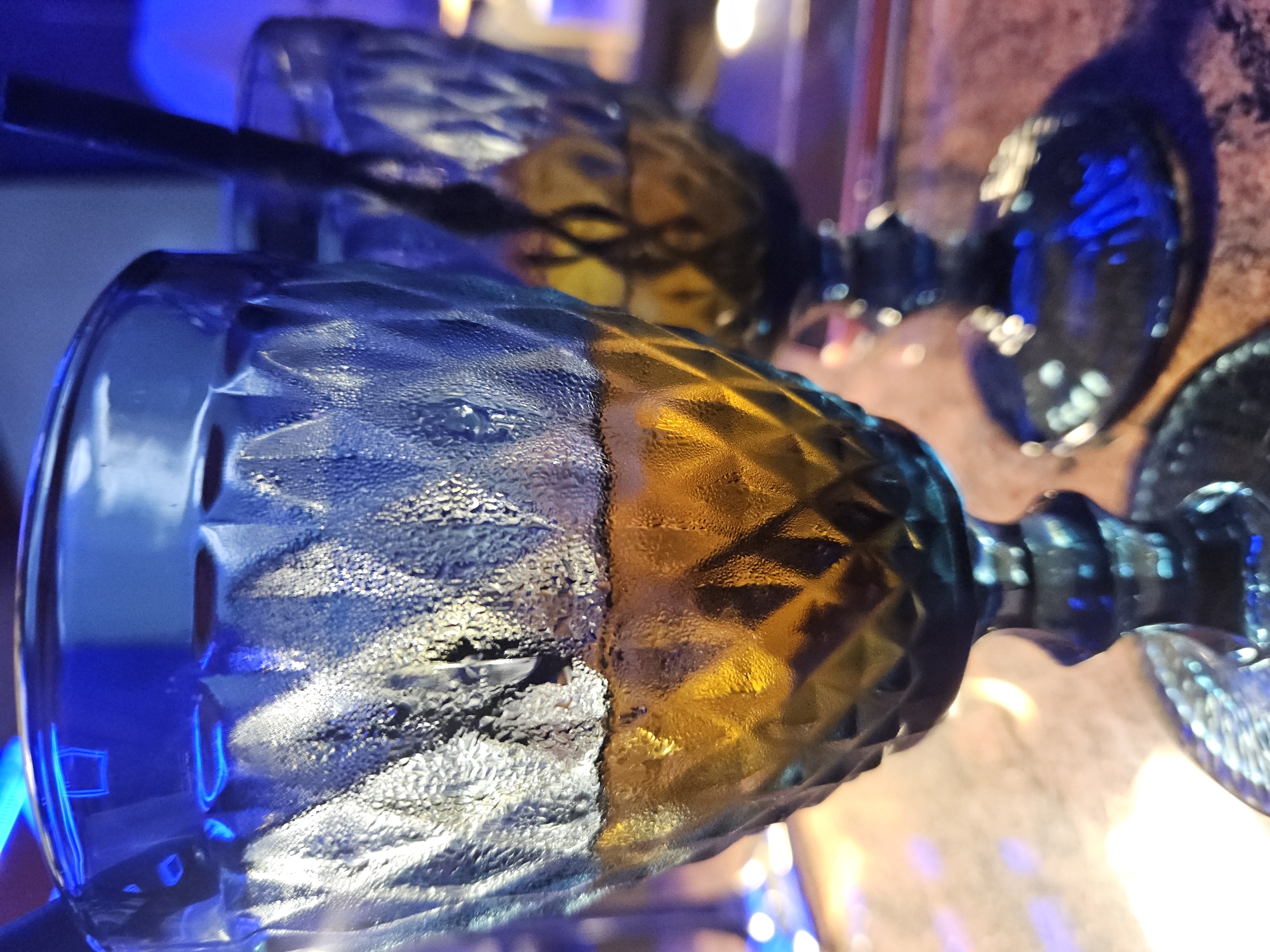 Two chilled stemmed glasses sit side-by-side on a reflective surface. The glasses are faceted, with a blue tint on one side and a yellow tint on the other. One glass has a black straw inside. Condensation is visible on the surface of the glasses. The location is Unknown, Unknown.