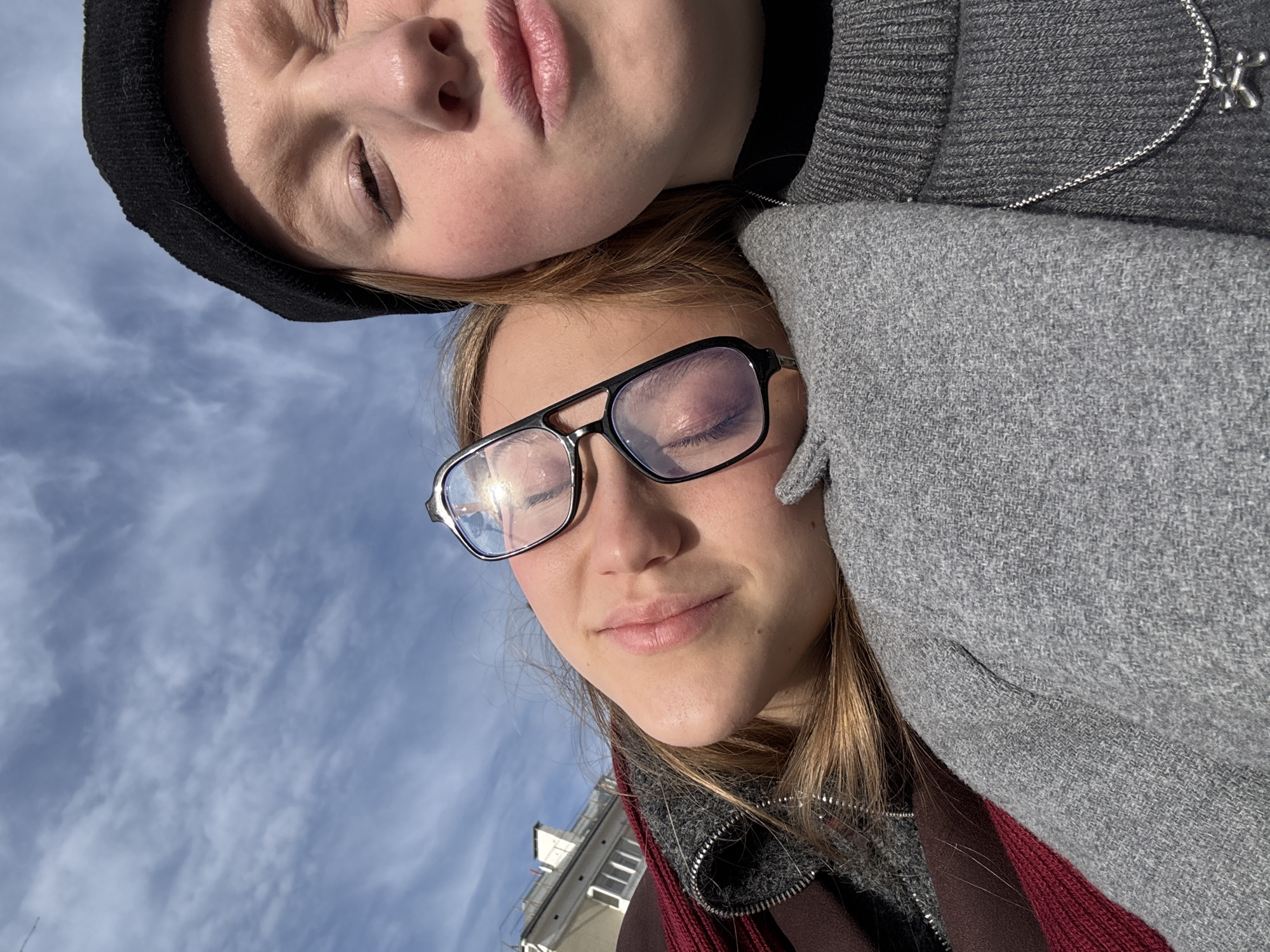 The image depicts two fair-skinned individuals in an outdoor setting. One individual is wearing eyeglasses and a grey overcoat with a red scarf beneath. The other individual is wearing a black beanie and a grey sweater, with a silver necklace visible. Both individuals have their eyes closed. The background includes a light blue sky with scattered clouds, and a building with multiple windows. Given the building and architectural style, it is plausible the location is in the city of Perm, Russia. The overall image appears to be a selfie taken from a low angle.