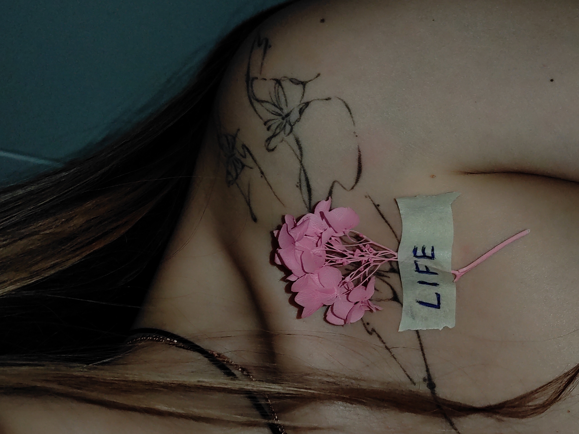 A close-up shot focuses on a person's upper chest and shoulder area. A tattoo of floral outlines is visible on the upper shoulder. A small pink flower is taped to the skin, the tape bearing the word "LIFE" written in blue. A delicate gold necklace adorns the neck. Strands of brown hair frame the subject. The environment is a plain, likely indoor setting, as suggested by the solid color background, but no distinct features indicate the location in Chaikovsky, Russia. No actions or interactions are taking place.