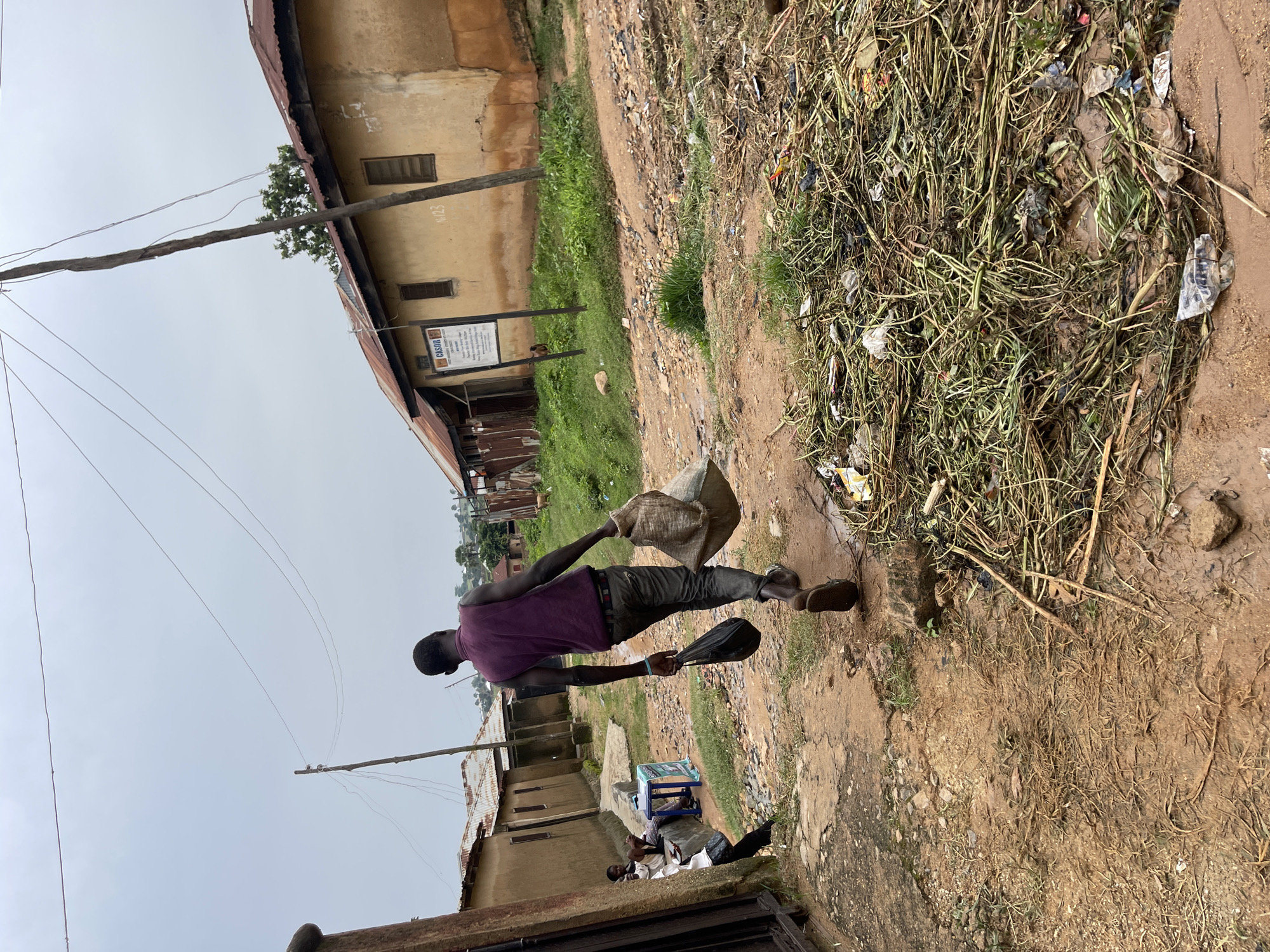 A man, wearing a purple shirt and dark pants, walks to the right of the frame carrying a burlap sack over his right shoulder and a black plastic bag in his left hand. He is walking on a dirt path in what appears to be a rural or semi-urban area. To his left, there are a few buildings with corrugated metal roofs and tan-colored walls. A sign with some writing on it is visible on one of the buildings. To his right, there is a pile of dry grass and debris, with some plastic trash mixed in. The ground is muddy and uneven. The sky is overcast, suggesting it is a cloudy day. There are power lines overhead. The scene appears to be in Jos, Nigeria. The man's posture and the way he carries the bags suggest he might be transporting goods or waste. The overall impression is one of a mundane daily activity in a modest setting.