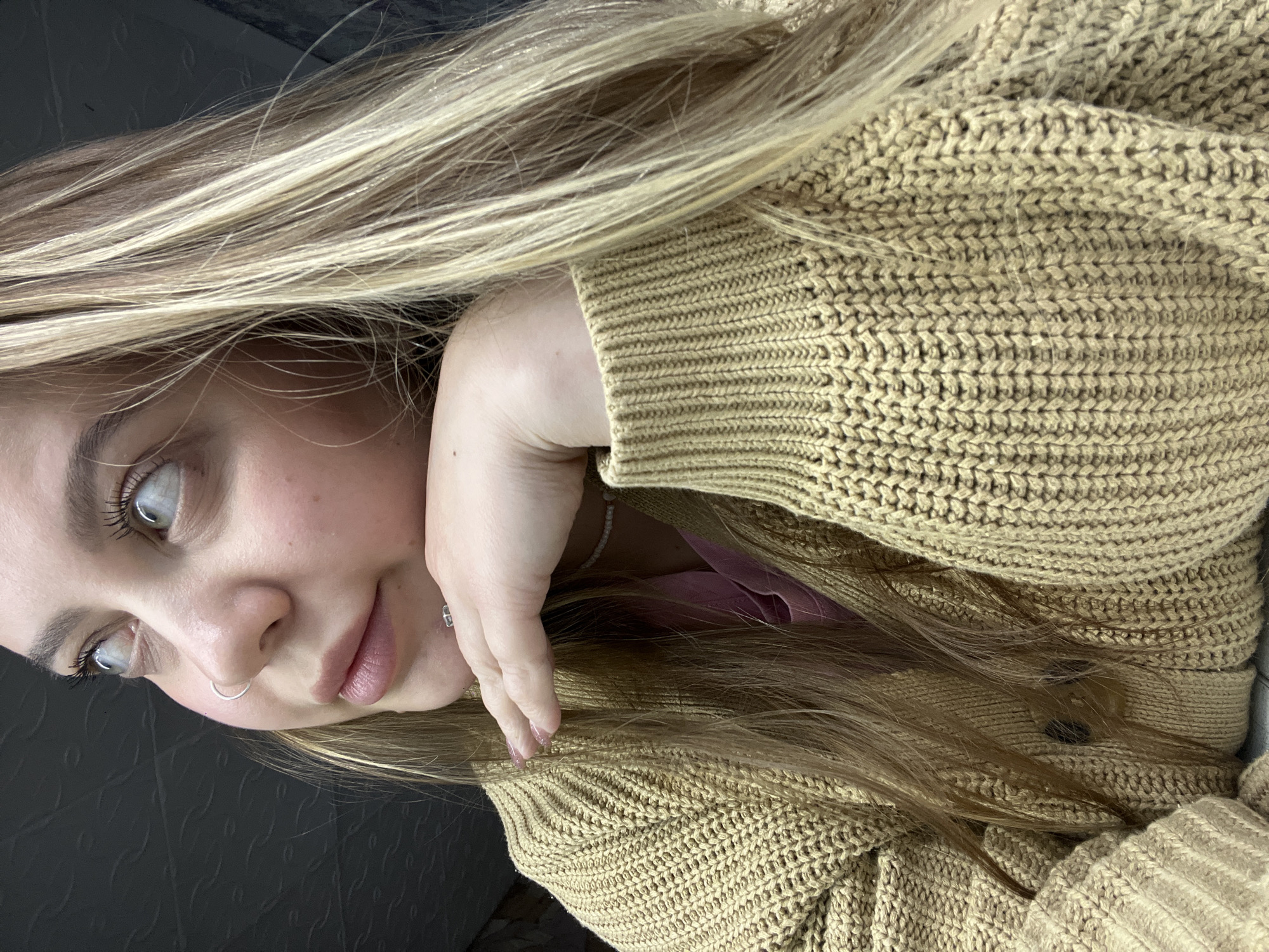 A close-up, vertically oriented portrait captures a young woman, likely indoors. Her head and upper torso are visible, positioned at an angle. She has long, straight hair with a mix of blonde and light brown strands, styled to fall over her shoulders and part of her outfit. She is wearing a light tan or camel-colored chunky knit cardigan or sweater.
The woman's left hand is gently placed near her chin, with her arm bent, creating a relaxed pose. Her face shows light-colored eyes, possibly blue or grey, which are directed slightly upwards and to her left (the viewer's right). She has prominent eyelashes, and her lips are slightly parted, conveying a neutral or serene expression. Notable accessories include a silver septum nose ring and a small silver stud earring in her visible right earlobe. A delicate, light-colored bracelet is also visible on her left wrist.
The background is dark, appearing to be an interior setting, possibly a car or a room with a dark wall, as faint textured patterns are discernible. The lighting on her face and hair suggests natural light, indicating daytime. The image has a shallow depth of field, bringing her features and the texture of her sweater into sharp focus. No text is visible in the image. The provided location context is Novopokrovskaya, Russia.