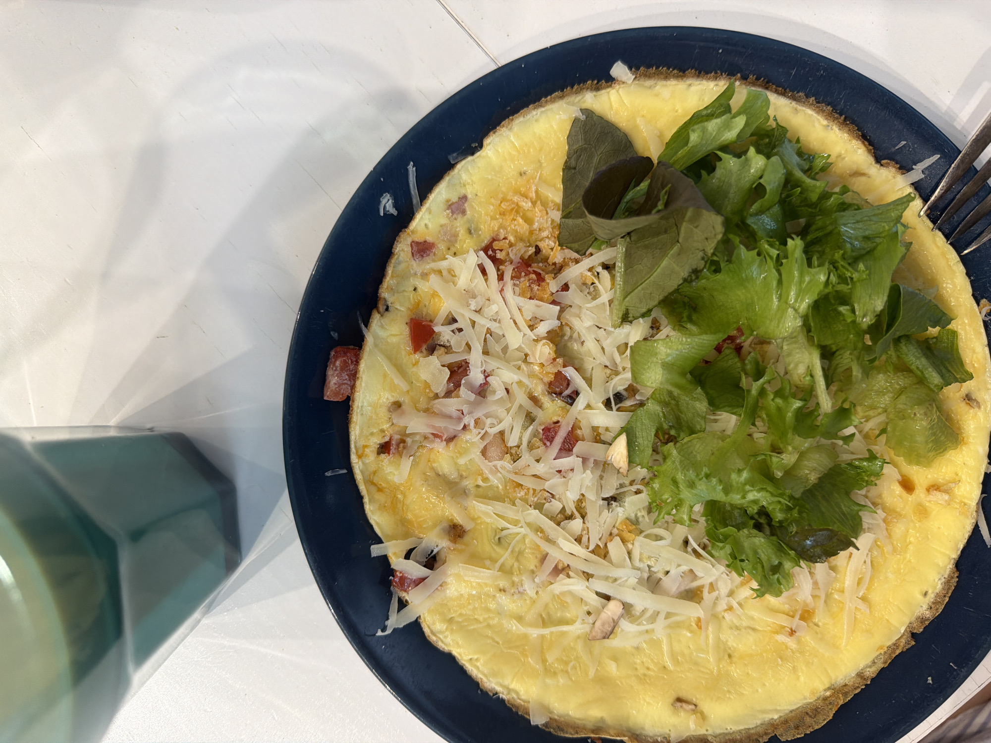 The image displays a meal set on a white, possibly wooden, surface. A dark blue, round plate occupies the lower and central portions of the frame, holding a prepared omelette or frittata. The omelette is a solid yellow, with slightly darker, crisped edges. It is topped with a generous layer of shredded white cheese, numerous small diced red components (consistent with bell peppers or tomatoes), and scattered light brown fragments (possibly mushrooms or nuts). A substantial portion of fresh green leafy salad, comprising various lettuce types, is placed on one side of the omelette. In the upper left quadrant, a faceted, transparent glass or bottle containing a light green liquid is partially visible. A silver-colored metal fork rests on the lower right edge of the plate, its tines pointing towards the food. The background surface is uniformly white with subtle linear textures. The specific location within the city of Chaikovsky, Russia, cannot be determined from the visual information provided. No individuals are present.