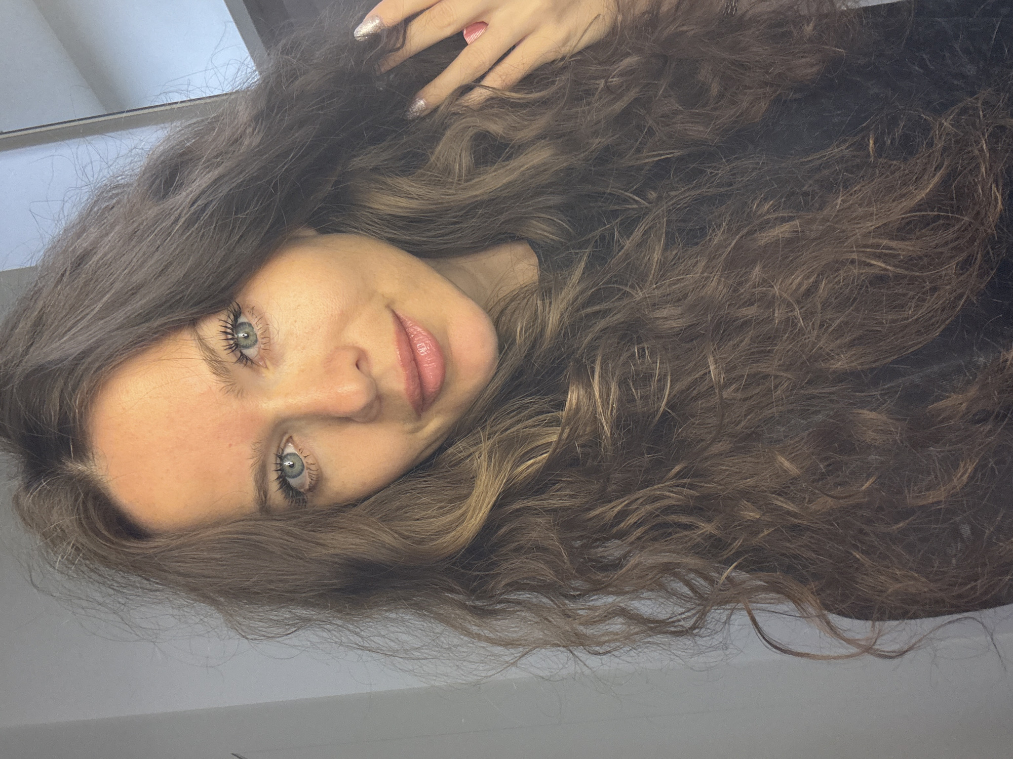 The image is a medium shot selfie of a woman with long, wavy brown hair. She has blue eyes, light skin with freckles, and is wearing subtle makeup. Her lips are slightly parted, and she is looking directly at the camera. Her right hand is raised to her hair, revealing a sparkly manicure and a pink ring. She is wearing a dark colored top. The background is a plain, light-colored wall and a reflective surface, possibly a mirror, which shows a bit of the ceiling. The lighting suggests an indoor environment. There is no discernible location within the city of Khimki, Russia, visible in the image.