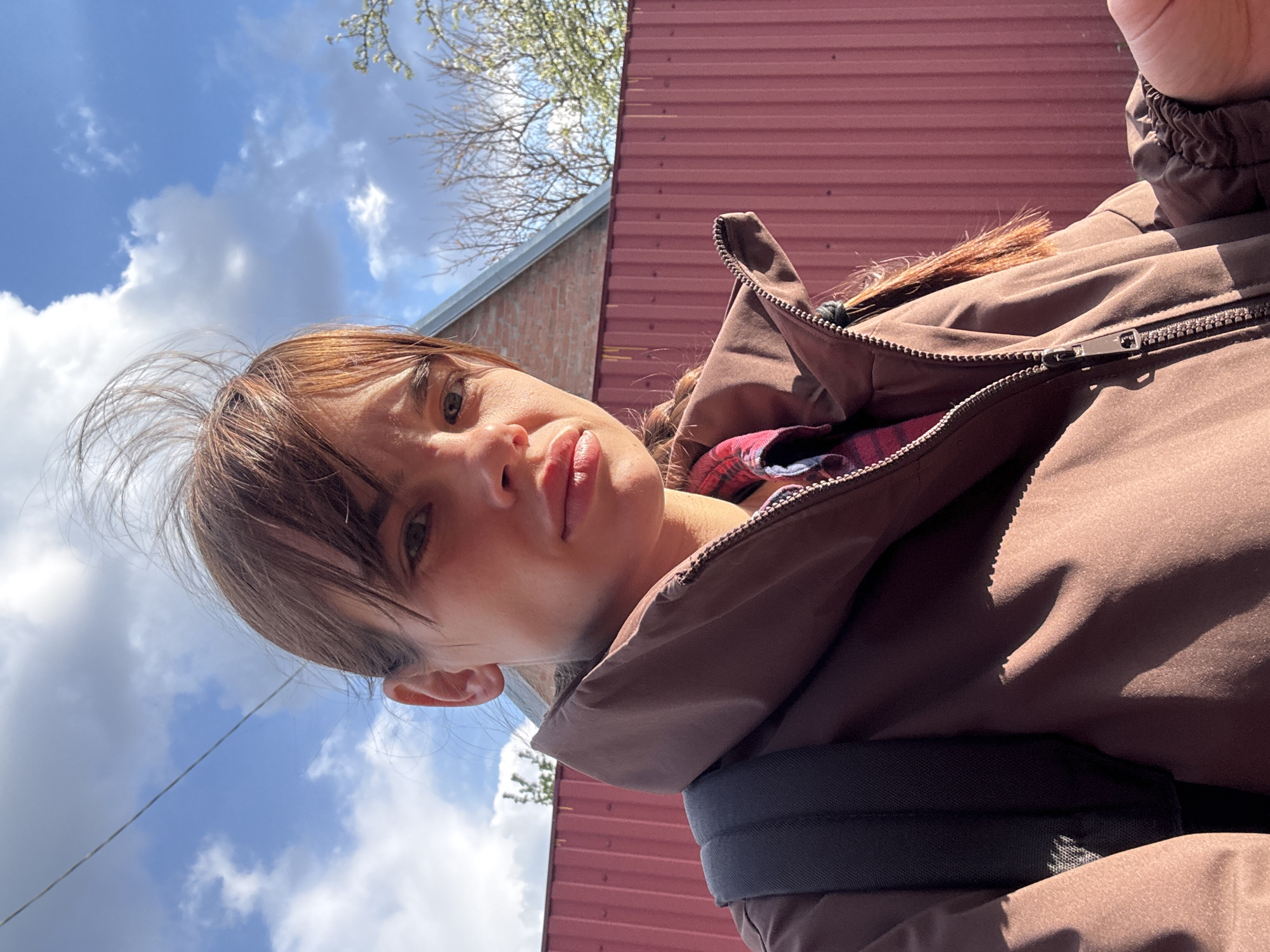An outdoor selfie, likely taken in Rostov-on-Don, Russia, features a young person looking directly into the camera from a slightly low-angle perspective. The person has brown hair with bangs, some strands appearing wind-blown on one side. Their expression is neutral or slightly contemplative. They are wearing a brown or dark beige puffer jacket, partially unzipped, revealing a red and black plaid shirt underneath. A small braid of reddish-brown hair is visible near the jacket's zipper, and a black strap, possibly from a backpack, is visible on their shoulder.
The setting is outdoors on a bright, sunny day with a clear blue sky dotted with white cumulus clouds. Bare tree branches, possibly with early buds, are visible against the sky, suggesting springtime. In the background, there is a prominent red corrugated metal wall or roof of a building, and to its left, a section of a light brown brick wall. Strong sunlight casts distinct shadows across the scene and the person's jacket. No visible text is present in the image.