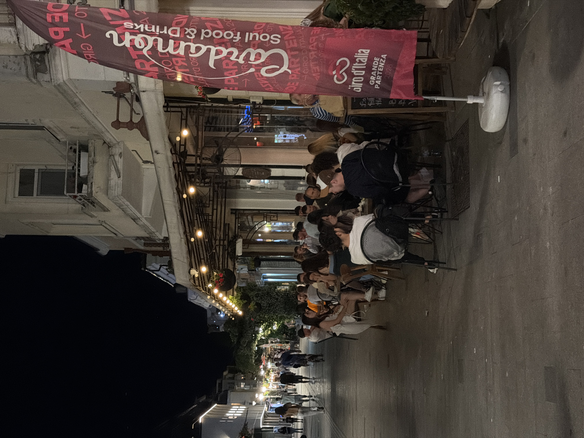 The image shows a street scene at night, likely in Burgas, Bulgaria. A long, maroon banner with white text hangs across the foreground, displaying "Cardamon Soul food & Drinks" and "Giro d'Italia GRANDE PARTENZA 2026". This suggests a street-side dining establishment or event.
Several people are seated at tables outside, engaged in conversation and dining. The lighting is warm, emanating from string lights overhead and interior lights from the buildings. The street itself is paved, and there are plants and trees visible, suggesting a lively urban environment. Further down the street, more people are walking, and lights from other establishments illuminate the scene. The overall atmosphere appears relaxed and social, typical of a pleasant evening outdoors. The presence of the banner and the outdoor seating indicates a public space designed for leisure and consumption.