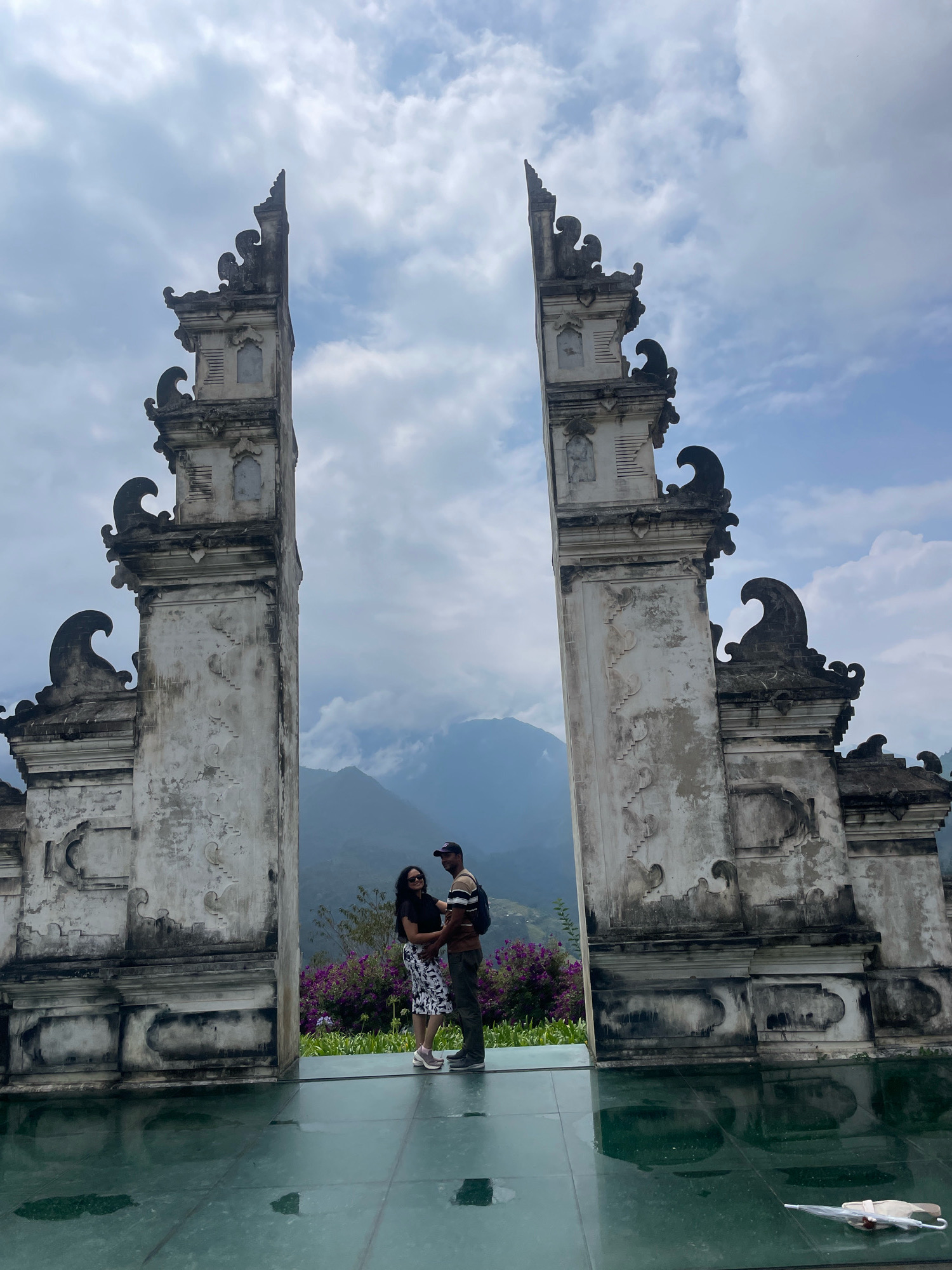 A couple stands embracing between two ornate, weathered stone gates at a scenic overlook in Sa Pa Ward, Vietnam. The gates, featuring intricate carvings and pagoda-like roofs, frame a view of misty mountains in the background. The couple is positioned on a reflective glass surface, which mirrors the sky and the surrounding landscape. The woman wears a black top and a patterned skirt, while the man wears a striped t-shirt, a baseball cap, and dark pants. A child is held by the man. Purple flowers and lush greenery are visible to the right of the couple. The sky is partly cloudy. An umbrella lies open on the glass surface in the foreground.