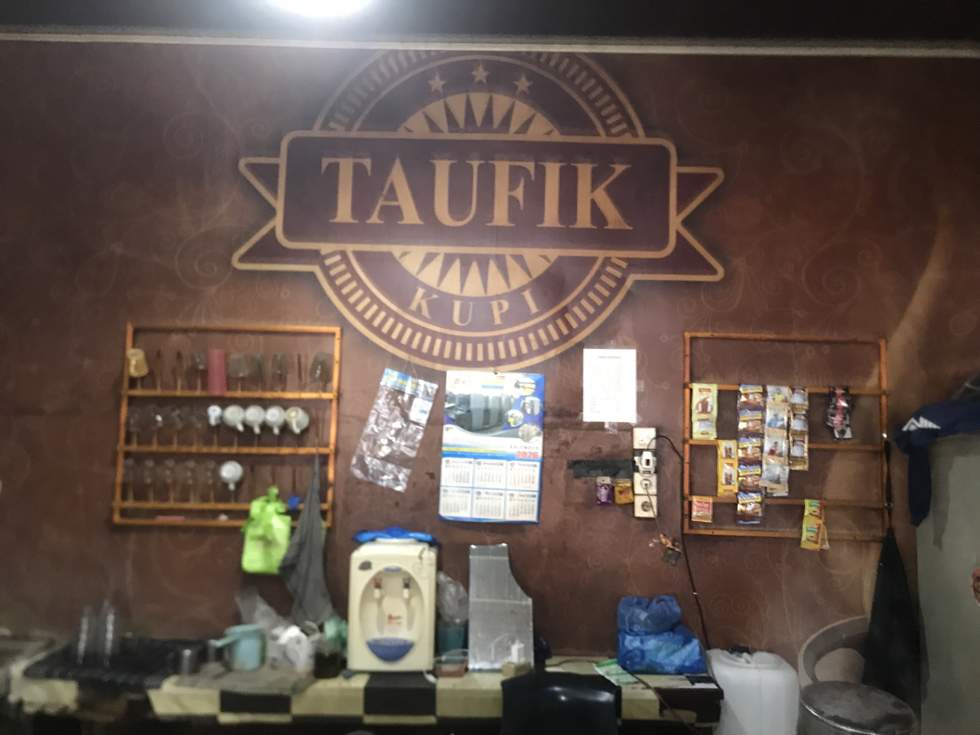 The image depicts a cluttered interior space, possibly a small shop or food stall, in Lhokseumawe, Indonesia. The wall features a brown patterned wallpaper with a large logo displaying "TAUFIK KUPI." Below the logo, a counter is covered with a black and white checkered pattern. Various items are placed on the counter, including a water dispenser, a plastic file organizer, a blue plastic bag, and what appear to be cleaning supplies. Shelves on either side of the wall hold drinking glasses and small packets of food or beverages. A calendar dated 2026 is also visible on the wall. The lighting in the space is dim, with a bright light source illuminating the upper portion of the image.