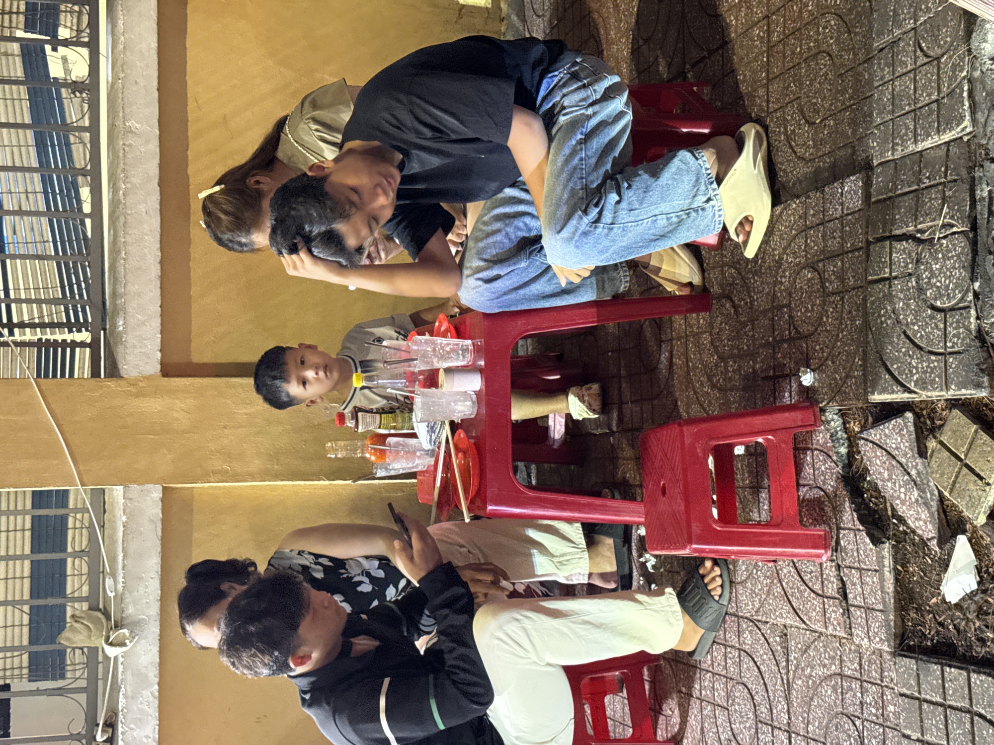 The image depicts a group of people, including two young boys, gathered around a low red plastic table on a tiled outdoor area. The setting appears to be a casual dining spot in Nha Trang, Vietnam, possibly a street food stall or small eatery, as suggested by the outdoor seating and the type of furniture. The people are seated on red plastic stools. On the table are several glasses, bottles of condiments, and red plates with chopsticks. One of the young boys is looking towards the camera with a serious expression, while the other is looking down at his phone. The background shows a building with yellow walls and a metal railing. The ground is paved with patterned tiles, some of which are broken and appear to be undergoing repair or showing disrepair. The lighting suggests it is evening or nighttime.