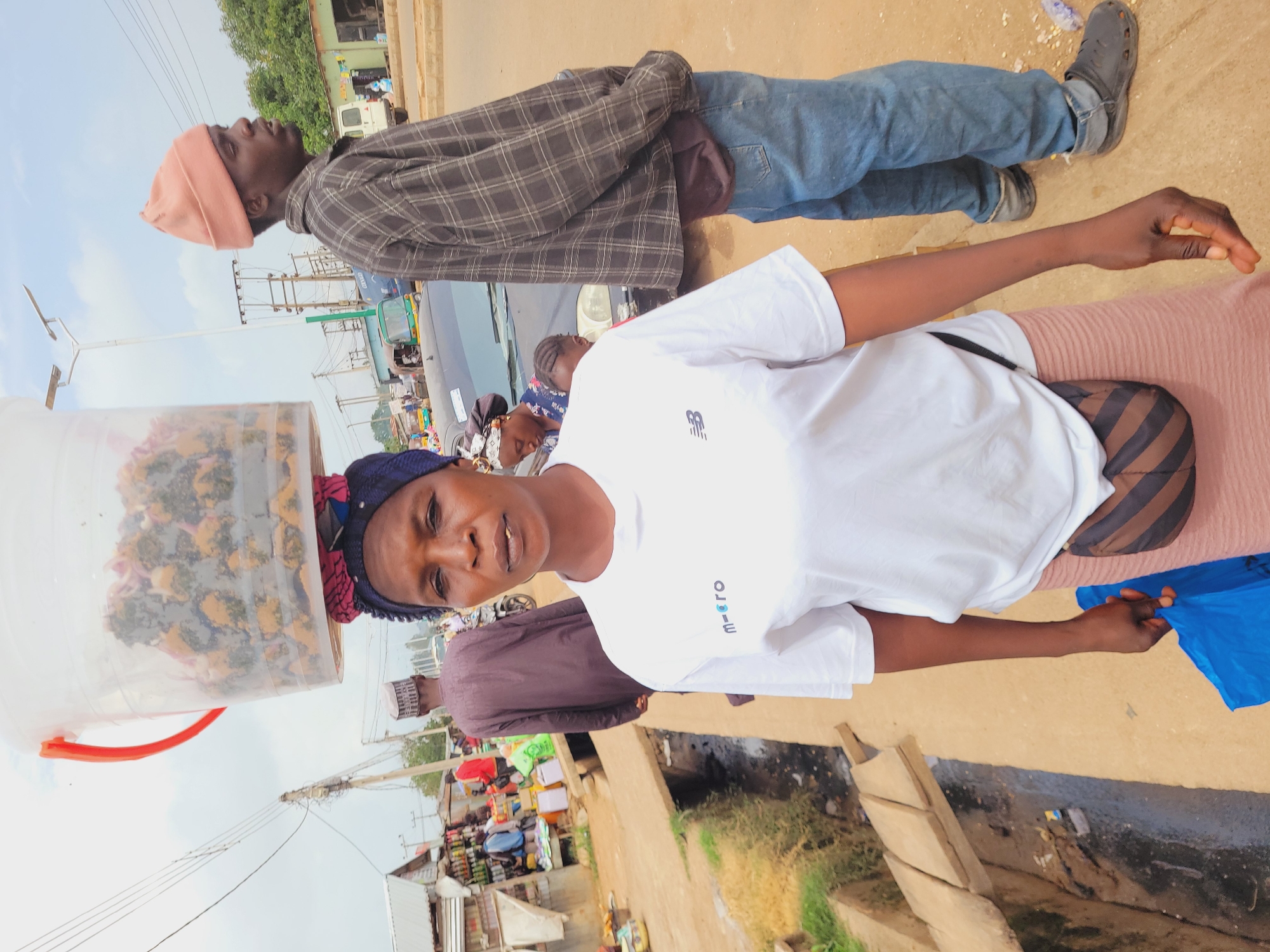 A medium shot captures a Black woman in Bukuru, Nigeria, balancing a large, clear plastic bucket filled with what appears to be food items on her head. She is wearing a white t-shirt with a "m=ero" logo and a light pink skirt, along with a brown and black striped fanny pack. A blue plastic bag is in her left hand. To her right, a Black man wearing a peach beanie, a plaid shirt, and blue jeans stands facing right, looking away from the camera. The background shows a dusty street with market stalls, a ditch filled with dark water and debris, and various vehicles, including a black car, a tuk-tuk, and motorcycles. Overhead power lines and streetlights are also visible. The overall scene depicts a typical street market environment in the city.