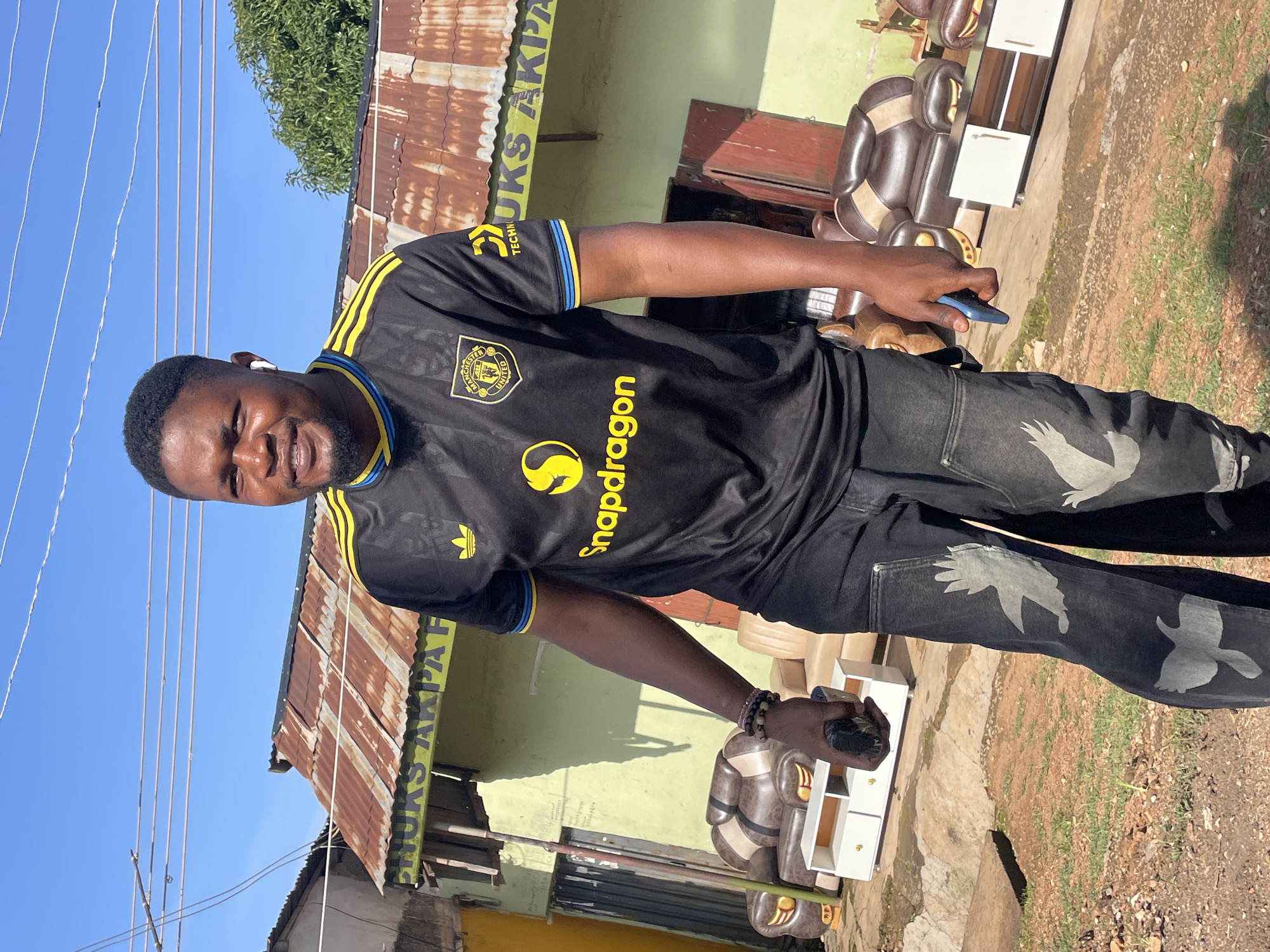 A medium shot captures a Black man standing outdoors in front of a small shop with a corrugated iron roof. The man, wearing a black and yellow Manchester United jersey and black jeans with white bird-like graphics, smiles as he looks at the camera. He has short, dark hair and a beard, and is wearing white wireless earbuds. In his right hand, he holds a dark-colored object, possibly a phone. In the background, to the left and right, are two sets of brown and cream-colored furniture, including sofas and a television stand. The shop itself is painted a pale green and has a yellow sign with text that is partially obscured but appears to read "CHUKS AKPA F". The sky is clear and blue, with a few power lines crossing the frame. The ground is dirt and patchy grass. The location is identified as Jos, Nigeria.