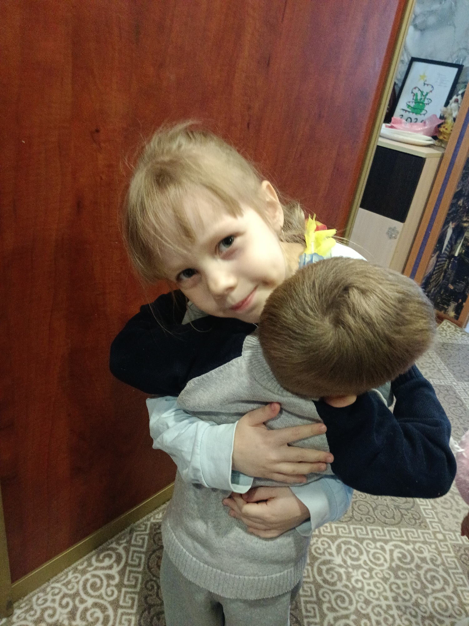 Two young children are captured indoors in Ольховка, Russia. In the foreground, a blonde-haired girl with bangs and light-colored eyes looks directly at the camera with a slight smile. She is wearing a light blue long-sleeved shirt under a grey sweater vest and has dark blue long sleeves extending from the vest. A yellow floral accessory is visible near her right ear. She is embracing a younger child, whose head, covered in short brown hair with a prominent crown swirl, is visible from the back. This second child appears to be wearing a dark blue long-sleeved top and grey pants. The background features a reddish-brown wooden door or wall paneling to the left and center. To the right, a narrow vertical surface, possibly a door frame or cabinet edge, reflects a portion of the room, revealing a framed picture with a white border depicting a green design and the year "2023," situated on a light-colored surface with a pink object. The floor is covered by a light-colored rug with a repeating pattern of stylized scrolls and geometric shapes.