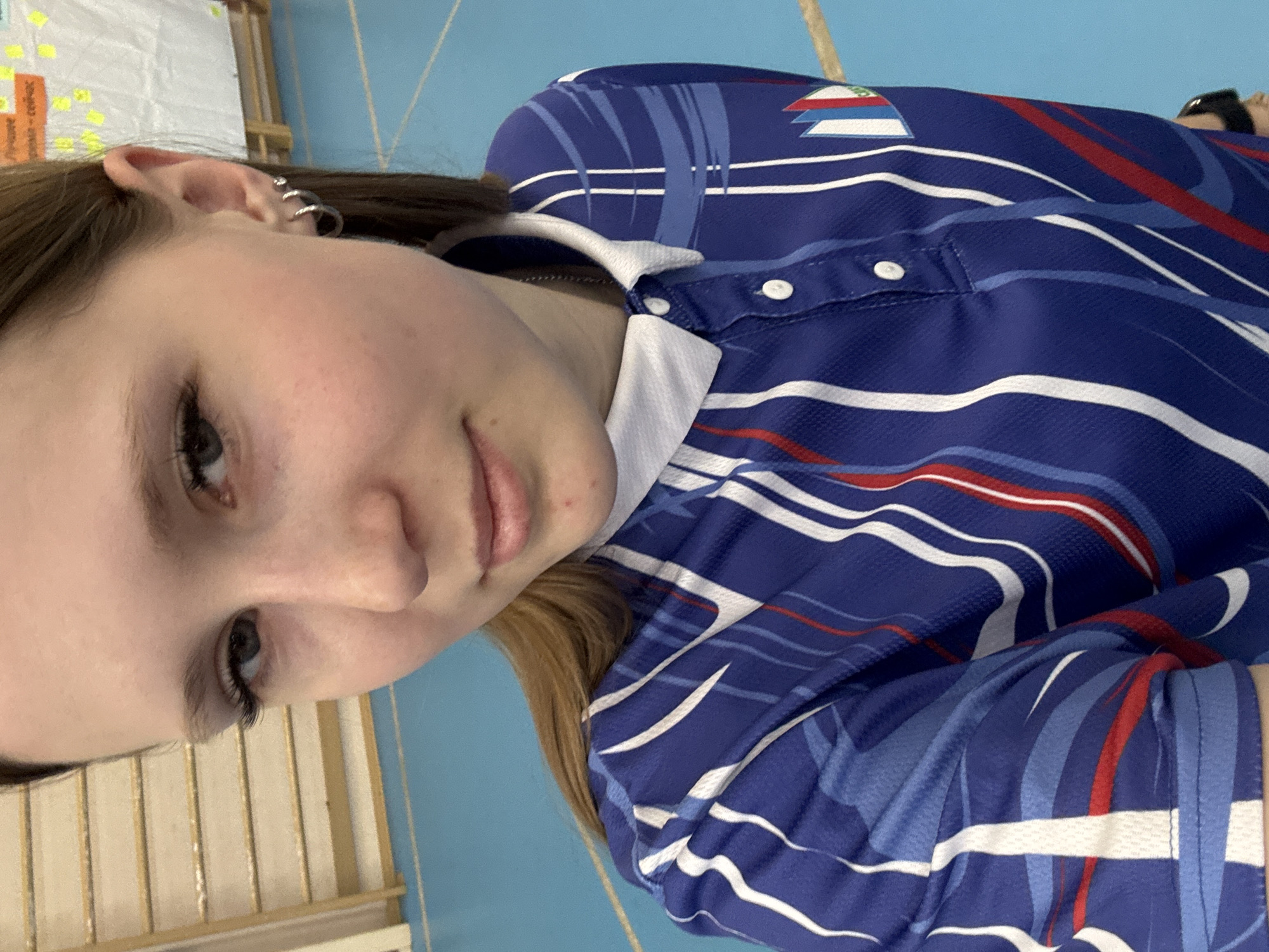 A young woman is the subject of this portrait. She has fair skin, brown hair, and her ear has multiple piercings. She is wearing a blue, white, and red striped collared shirt with a patch on the shoulder, with the flag of the country of Slovenia. The location appears to be indoors, with a blue wall and a white wooden structure visible in the background. Usolye, Russia is not evident in the image.