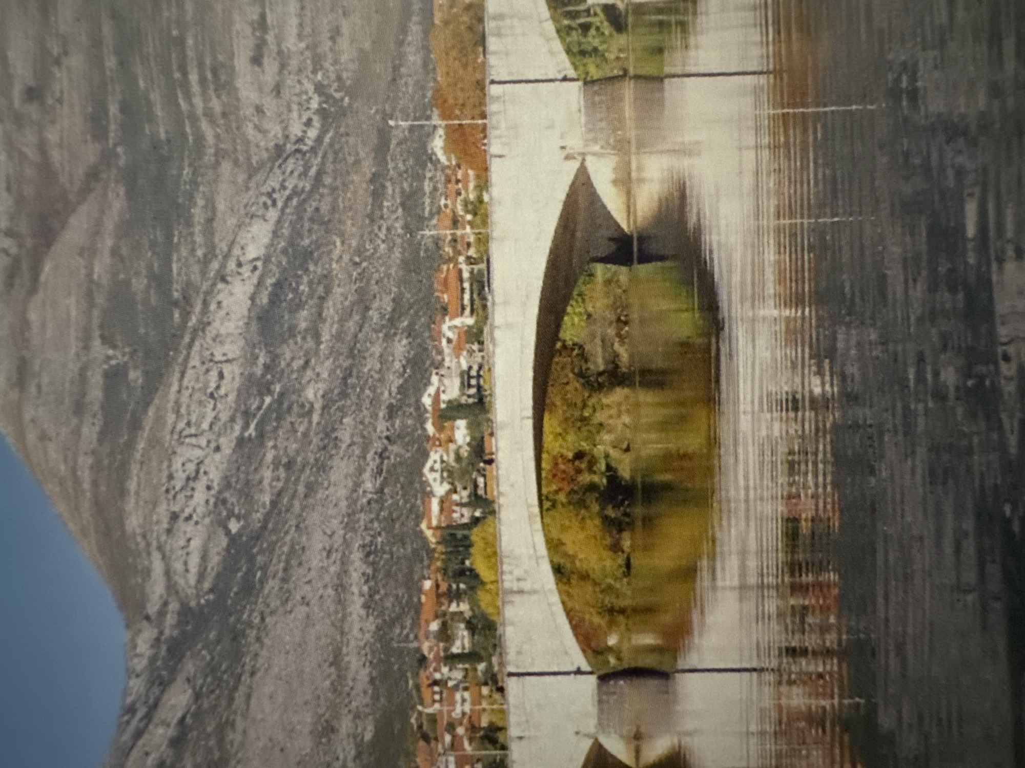 The image depicts a wide, arched concrete bridge spanning a body of water. The bridge is light in color and shows some signs of age. On the far bank, behind the bridge, a town with red-tiled roofs is visible, nestled against a backdrop of a rugged, barren mountain range. The mountain slopes are a muted gray and appear to be covered in rock and sparse vegetation. The sky above is a clear, bright blue. The water in the foreground reflects the bridge, the trees on the bank, and the town, creating a slightly distorted but colorful mirror image. The trees on the near bank, visible under the bridge, display autumn foliage in shades of yellow and green. The overall scene suggests a serene, possibly rural or small-town setting, with a strong natural landscape dominating the background.
