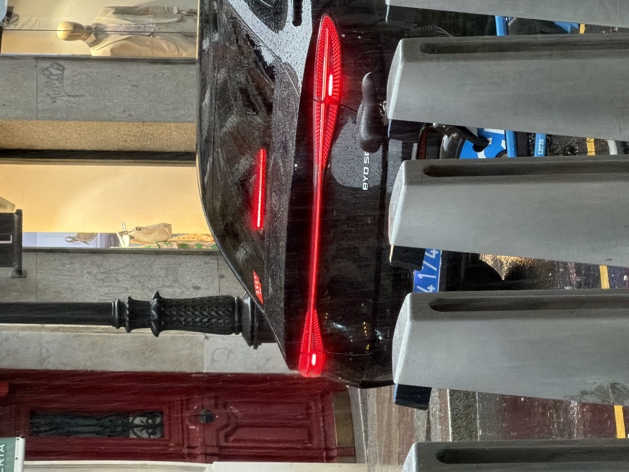 A black BYD Seal electric car is parked on a city street in Madrid, Spain, as suggested by the European license plate and architectural details. The car has distinctive vertical LED taillights. It is parked next to a series of light gray concrete bollards. Behind the car and bollards are urban elements, including a red door with decorative ironwork, a black wrought-iron lamppost, and a storefront window. A mannequin can be seen in the window of the store. The environment appears to be wet, possibly from recent rain.