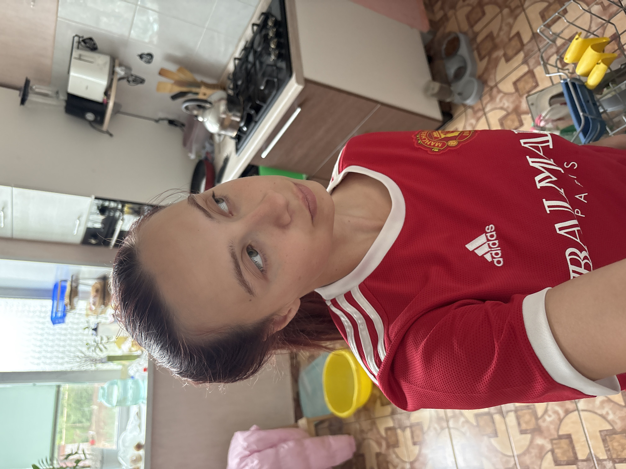 The image shows a young woman with dark hair pulled back, wearing a red Manchester United jersey with white stripes on the shoulders and white trim around the collar and sleeves. She is looking to her right, with her mouth slightly open. The background appears to be a kitchen. To the left, a window shows a blurred outdoor scene with greenery and a large blue bottle on the windowsill. A pink quilted item is also visible on the left. In the middle ground, a yellow bowl sits on the floor. To the right, a kitchen counter features a stovetop with a kettle and utensils. Above the counter, appliances including a toaster and a blender are mounted on a shelf. The floor is tiled with a patterned design. The overall impression is a candid shot taken within a domestic setting.