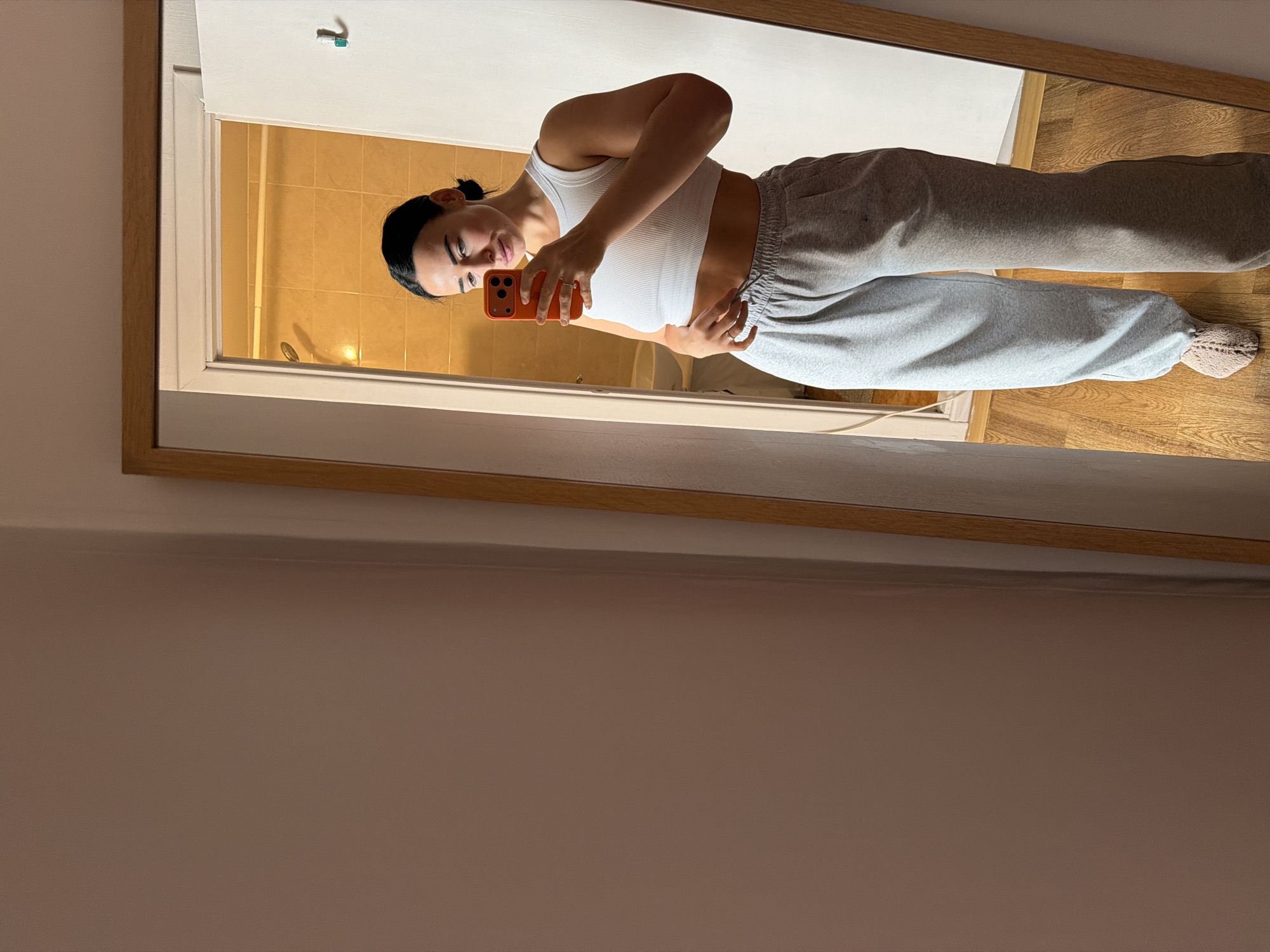 A young woman is captured in a full-length mirror selfie, with the image rotated 90 degrees clockwise. She stands casually, facing slightly to her right, holding an orange smartphone in her left hand to take the photo. Her right hand rests gently on her lower abdomen. She is wearing a white ribbed sleeveless crop top that reveals her midriff, paired with loose-fitting grey sweatpants and light-colored, possibly fluffy, slippers on her feet. Her dark hair is pulled back, and she offers a subtle, pleasant smile towards the phone's camera.
The mirror has a wooden frame and reflects a room behind the woman, which appears to be a bathroom or a tiled area with warm, amber-toned tiles on the walls and a shower fixture visible. A small white hook is also visible on the wall in the reflection. The floor in the reflected room is made of wood. The wall on the left side of the mirror, outside the reflection, is a plain light brown or beige color. The lighting in the scene is artificial and appears warm, contributing to a relaxed atmosphere. There is no visible text in the image.