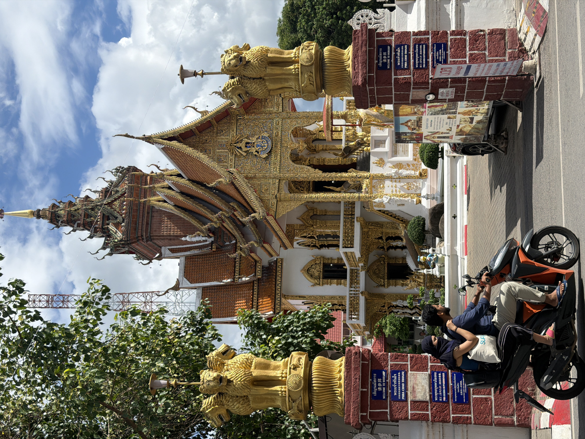 In the city of Chiang Mai, Thailand, a motorcycle carries two individuals, a man and a woman, as they approach the entrance of a temple. The man, in a blue shirt and light-colored pants, drives the motorcycle, while the woman, wearing a black headscarf and a blue top, sits behind him. The temple's architecture is characterized by its ornate golden decorations and multi-tiered roofs, topped with a golden spire. Flanking the entrance are two large golden statues of lions with multiple heads. Informational signs in Thai and English are posted on the brick pillars of the entrance. Behind the motorcycle, a small vehicle adorned with colorful images of temples is parked. The scene is set against a backdrop of lush green trees and a sky filled with white clouds.