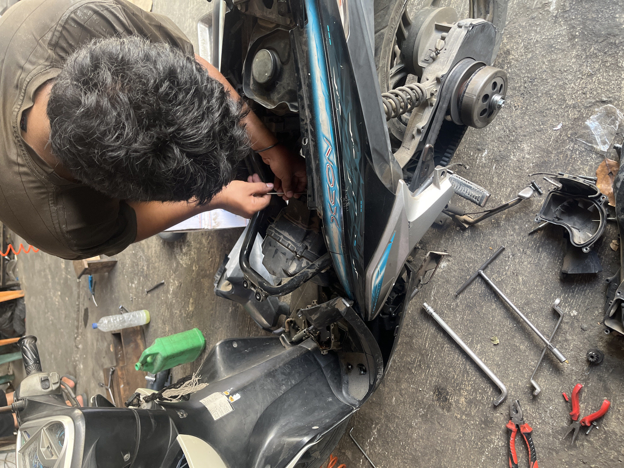 A man with dark, curly hair is working on a scooter in what appears to be a workshop or garage. He is wearing a dark brown t-shirt and is hunched over the scooter, with his hands engaged in adjusting or fixing a component near the engine. The scooter is partially disassembled, with panels removed to expose the internal mechanics. Various tools are scattered on the concrete floor around the scooter, including wrenches, pliers, and a screwdriver. A green plastic container and a bottle of clear liquid are also visible on the floor, suggesting maintenance or repair activities. The environment looks industrial with some items in the background that might suggest a busy workshop. The lighting appears to be natural and artificial, indicating an indoor setting. The overall scene depicts a mechanical repair in progress. Based on the provided hints, this image may be located in Banda Aceh, Indonesia.