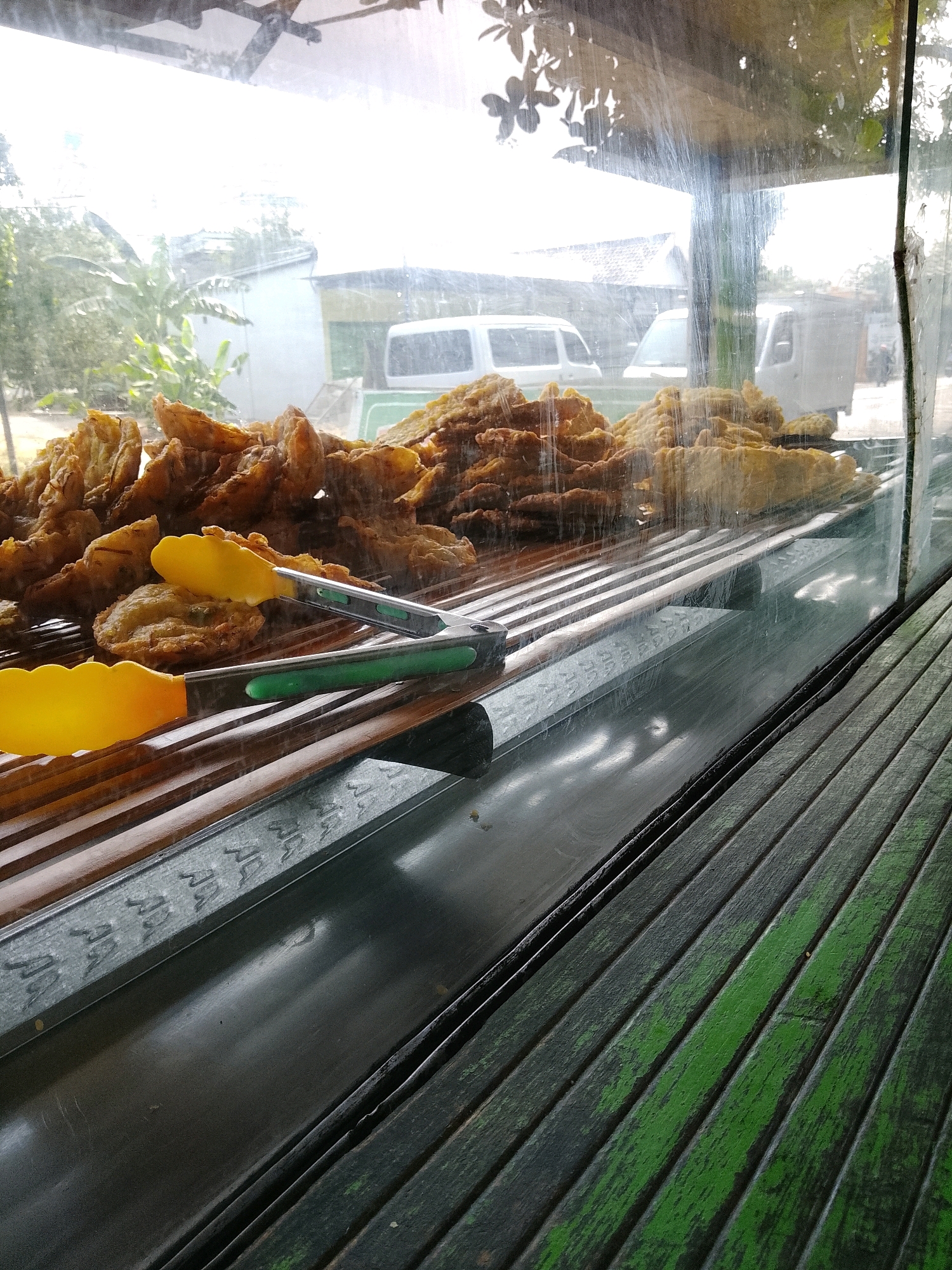 This image captures a close-up view of fried food items, likely bananas or plantains, displayed behind a clear plastic barrier, possibly a window or enclosure. Two yellow-handled tongs rest on a wooden slatted surface in front of the food. The background, visible through the barrier, shows an outdoor scene with trees, buildings, and what appear to be several white vehicles, including vans and a truck. The overall impression is that of a food stall or eatery, possibly in a street setting. The text overlays on the image suggest the location is Banaran, Indonesia. The lighting is bright, indicating daytime.