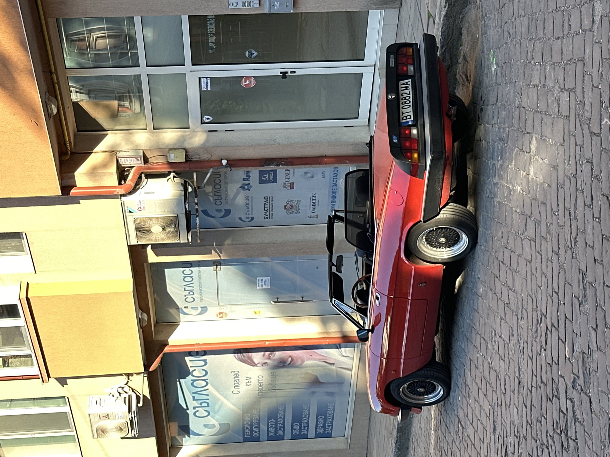 A red convertible Alfa Romeo Spider, model year 1983-1993, is parked on a cobblestone street in front of a modern building. The car, with its distinctive "Schnauzer nose" grille and pop-up headlights, has its license plate visible: BT 0882 MA. The building facade features large glass doors and windows, reflecting the surrounding environment. Multiple signs are visible on the building, indicating various insurance and financial services offered by "съгласие" (meaning "consent" or "agreement" in Bulgarian), including pension, life, general, and health insurance. One sign features a smiling woman, conveying a sense of optimism for the future. Air conditioning units are mounted on the building's exterior. The scene appears to be set during the daytime, with sunlight casting shadows and highlighting the textures of the cobblestones and building materials. The overall atmosphere is one of urban street life, with a classic car adding a touch of vintage charm to the modern setting.