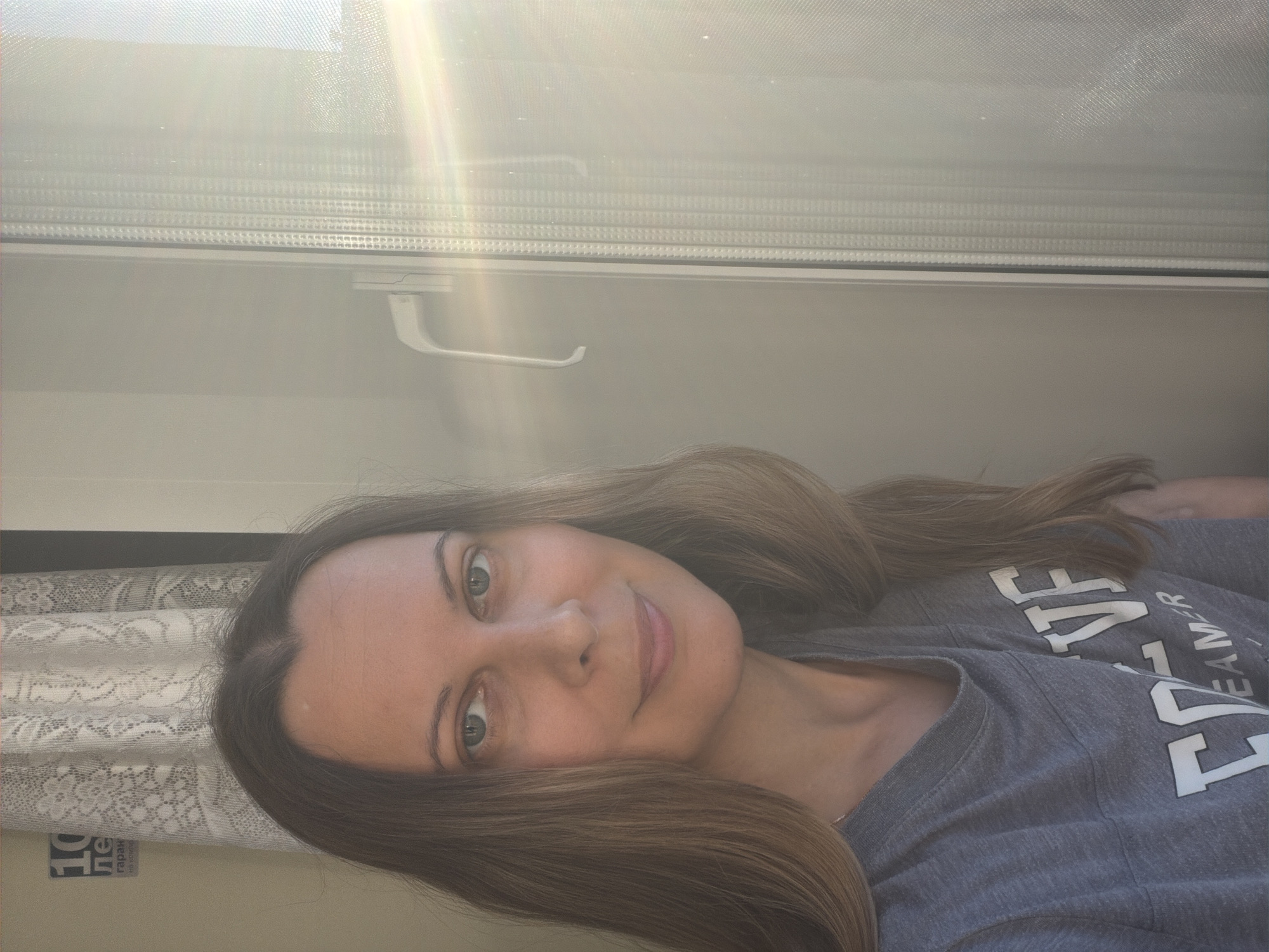 A Caucasian woman with long, light brown hair is positioned in the foreground, facing the camera and slightly to the left. She is wearing a grey t-shirt with white lettering that appears to say "CREATIVE DREAMER". She is standing in front of a window with vertical blinds. Sunlight is streaming in from the right side of the window, creating lens flare and illuminating the scene. A white window handle is visible on the right side of the frame. In the background, to the left of the woman, a portion of a light-colored wall is visible, with a white curtain hanging against it. A black and white sign with Russian text, possibly a business advertisement, is partially visible on the wall. The overall environment suggests an indoor setting, likely an apartment, with natural light. It is not possible to determine the specific location within the city of Izhevsk, Russia, based on the visual information alone.