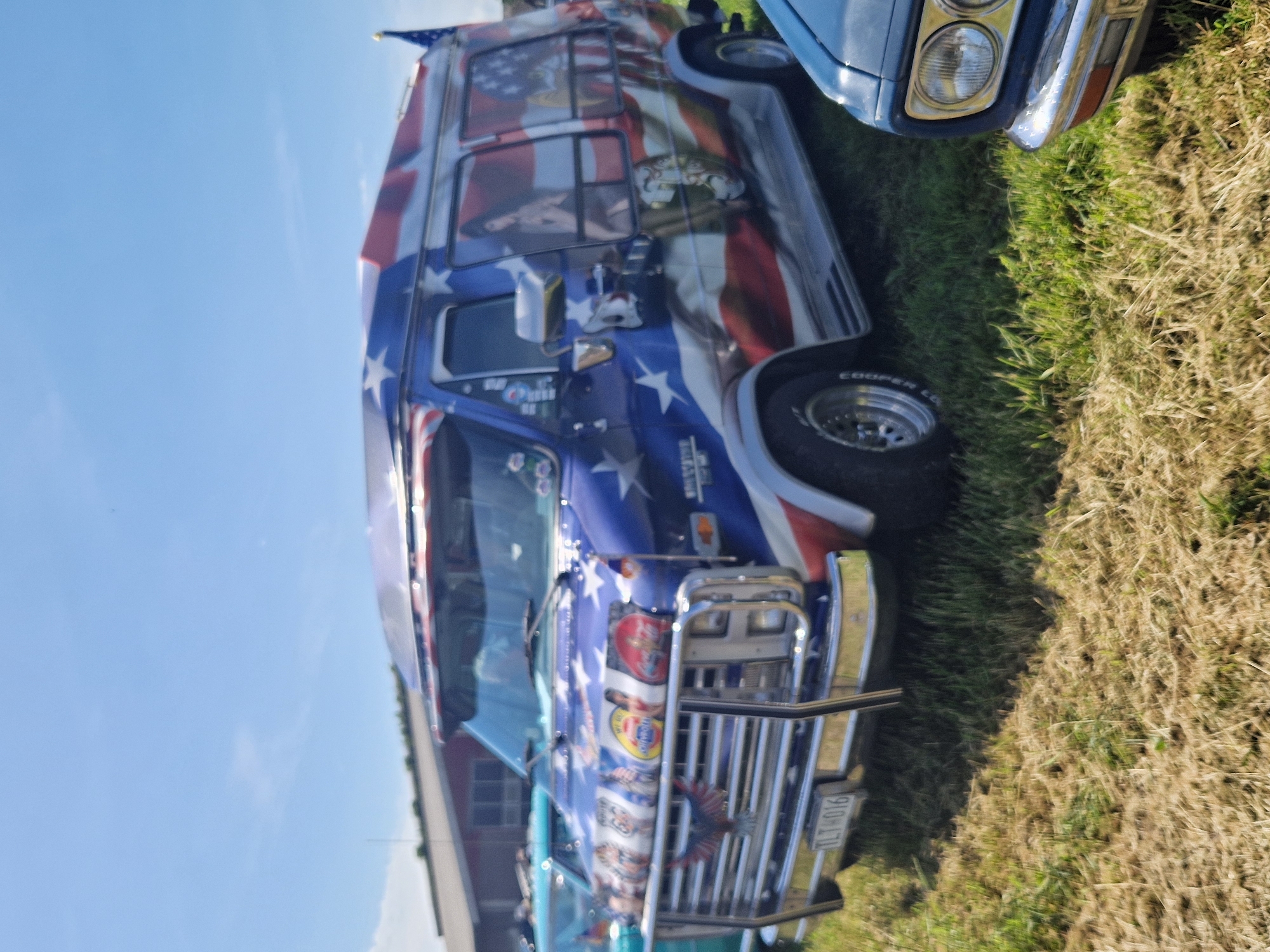 The image shows a van with an American flag and eagle motif parked on grassy ground under a clear blue sky. A portion of another vehicle, likely a car, is visible in the foreground on the right. The van has a chrome bumper and a license plate that reads "TLT 016". A small American flag is attached to the roof of the van. In the background, a building with a red roof is visible. The scene appears to be outdoors, possibly at an event or gathering. The location is identified as Edenberga, Sweden.