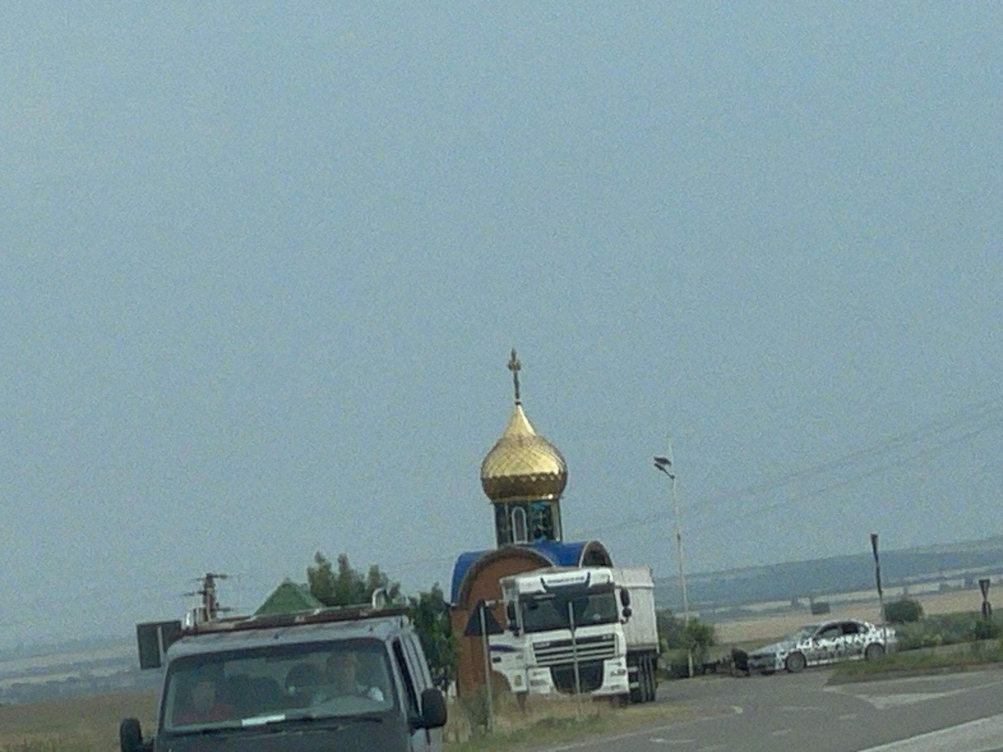 The media file depicts an outdoor scene with a clear, overcast sky suggesting a daytime setting. In the foreground, a dark-colored vehicle, possibly a van or SUV, is partially visible on the left, with people visible inside. Further to the right, a white truck is parked next to a small, domed structure that resembles a chapel or a roadside shrine. The dome is golden, and the structure has blue trim. Behind the truck and chapel, a silver car with a camouflage-like pattern is parked on the right side of the road. The background shows a rural landscape with fields and trees, indicative of a setting outside a major urban area. The overall impression is of vehicles stopped near a religious landmark along a road, possibly in a rural part of Congaz, Moldova, given the location context. There is no visible text in the media file.